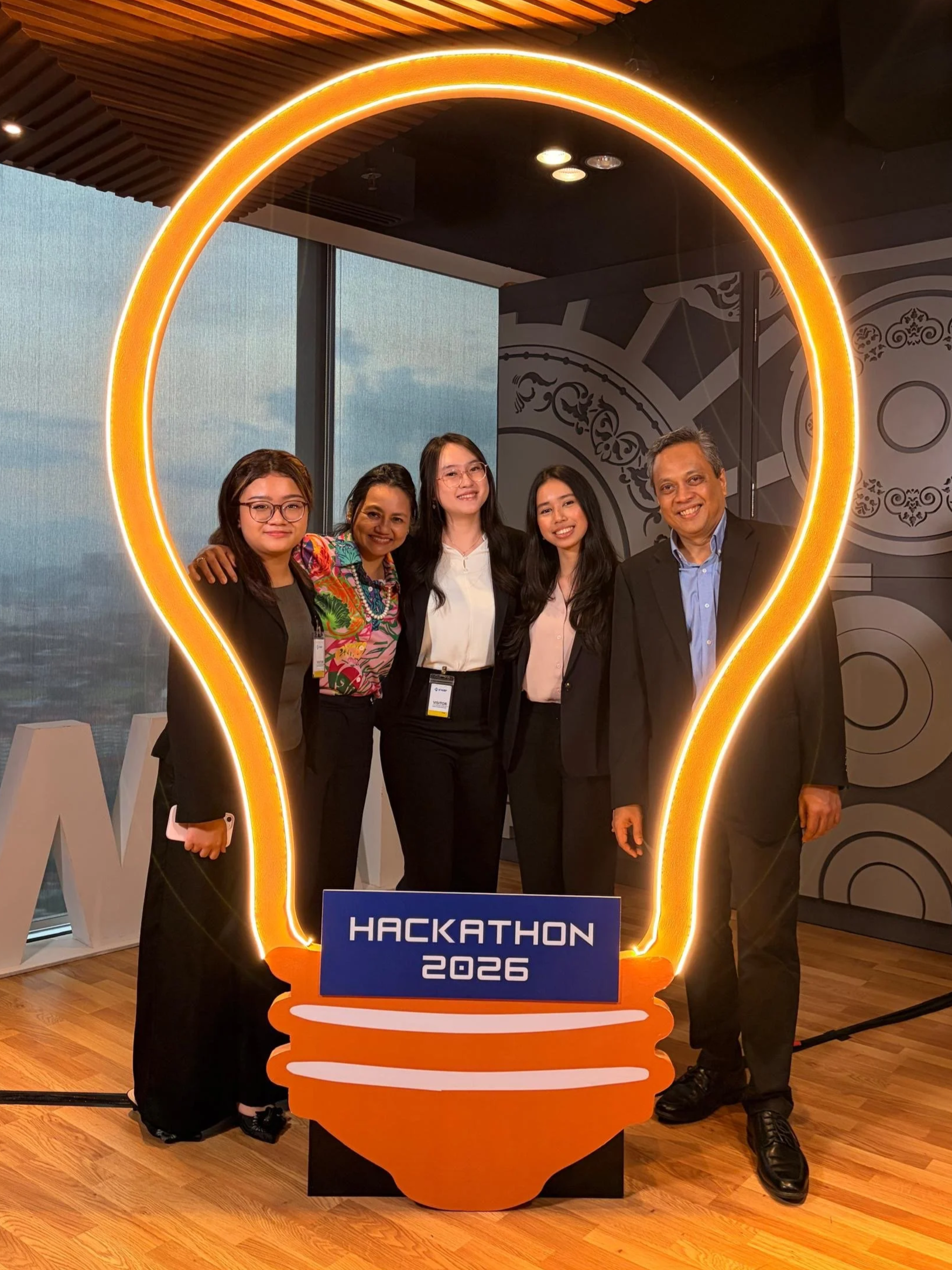 Group of five people posing behind a large illuminated lightbulb-shaped sign that reads "Hackathon 2026" at an indoor event.