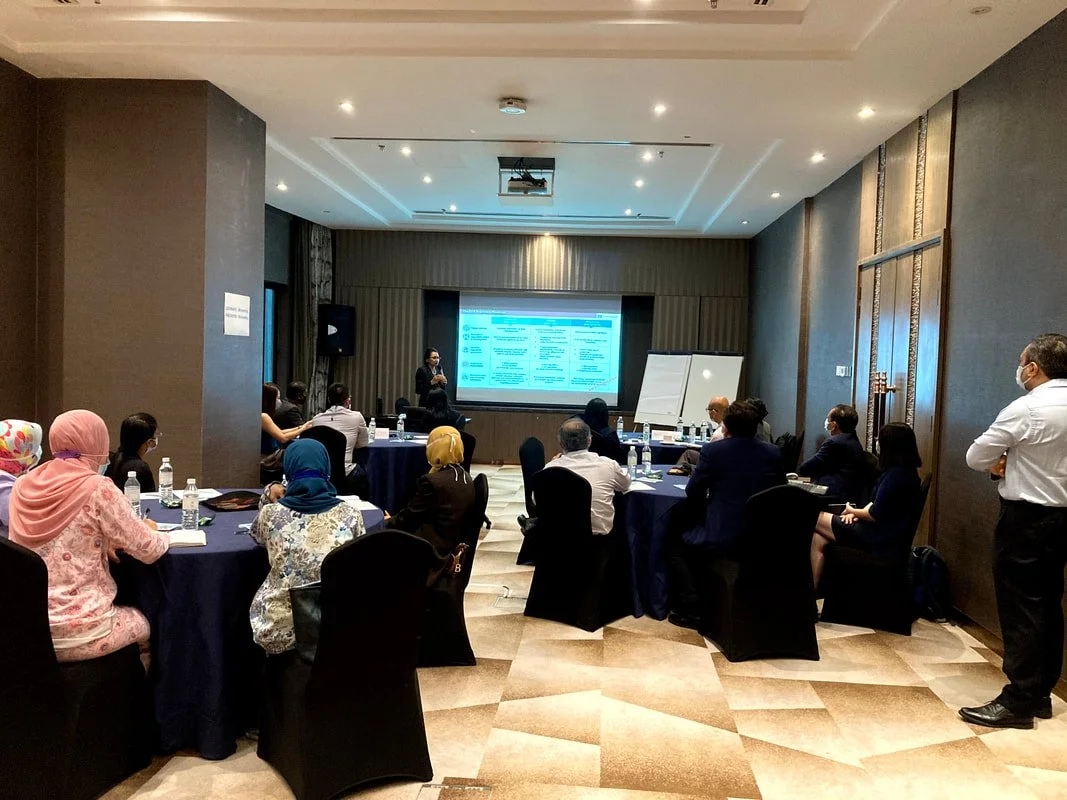 A business conference in a conference room with attendees seated at round tables, a presenter standing at the front near a presentation screen, some participants wearing headscarves, and bottles of water on the tables.