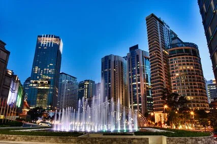 City skyline at dusk with illuminated skyscrapers and a fountain in the foreground