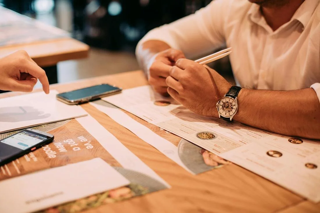 People sitting at a wooden table in a restaurant or cafe, looking at menus, with smartphones nearby.