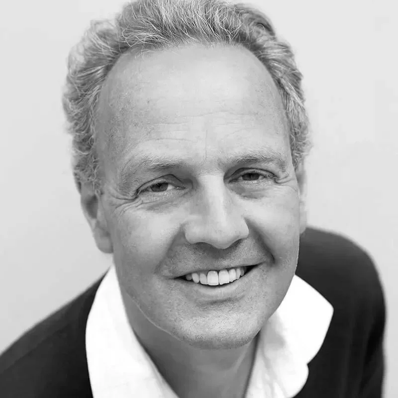 Black and white close-up portrait of a smiling middle-aged man with short, curly, gray hair, wearing a dark suit jacket and white shirt.