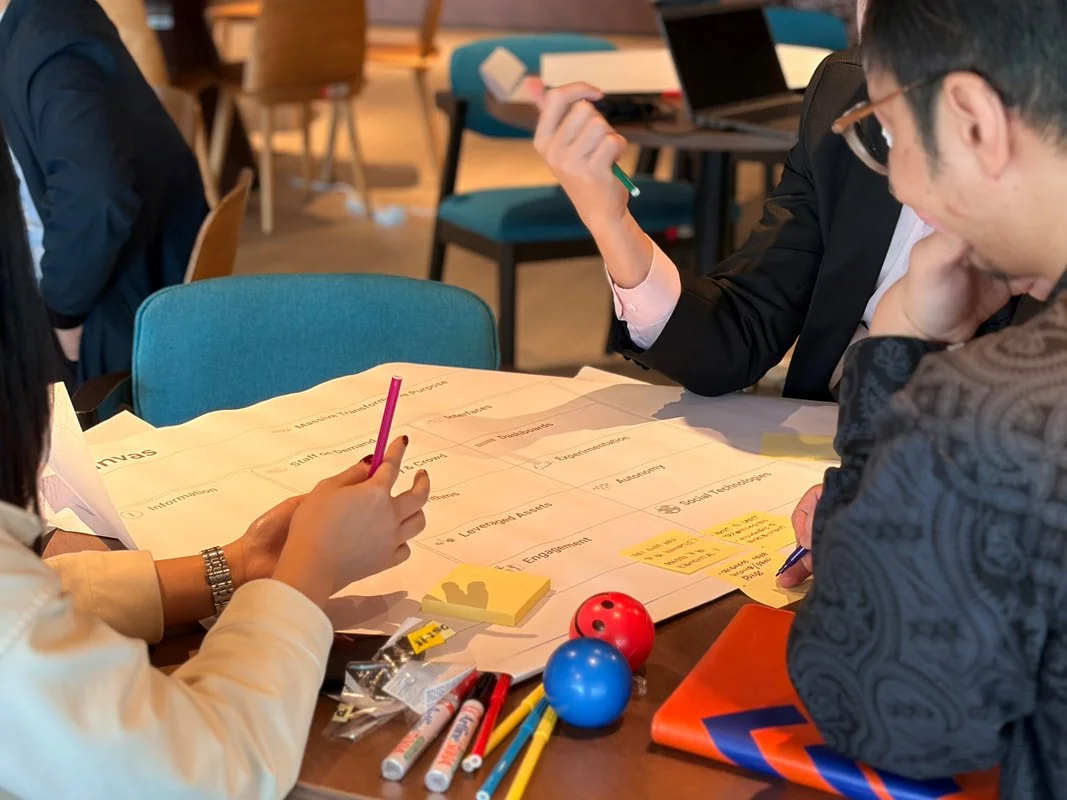Group of people working on a project at a table with charts, sticky notes, and colored balls, in a collaborative workspace.