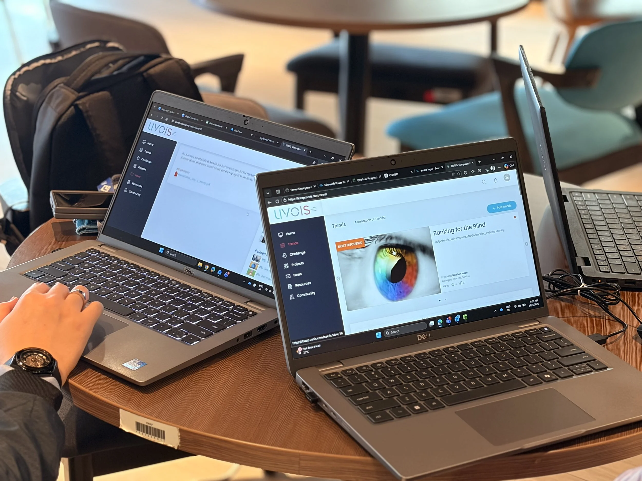 Multiple open laptops on a wooden table, displaying websites; a person's left hand is visible resting on one laptop, wearing a black watch, in a setting with chairs and tables in the background.