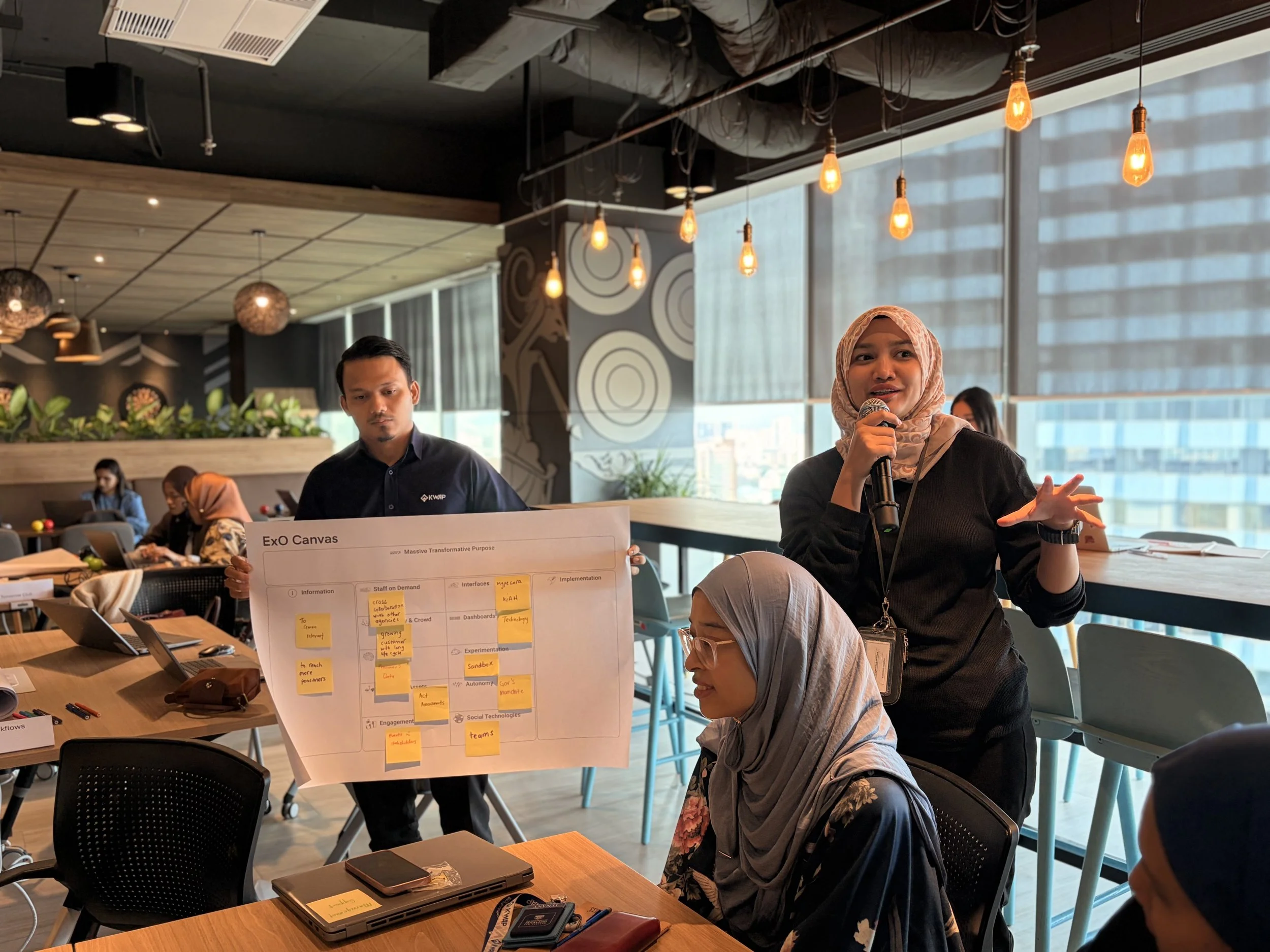 A woman wearing a headscarf and black outfit is speaking into a microphone while sitting at a table in a modern conference room with large windows. Other people are sitting nearby, and a presentation board with sticky notes is in front of her.