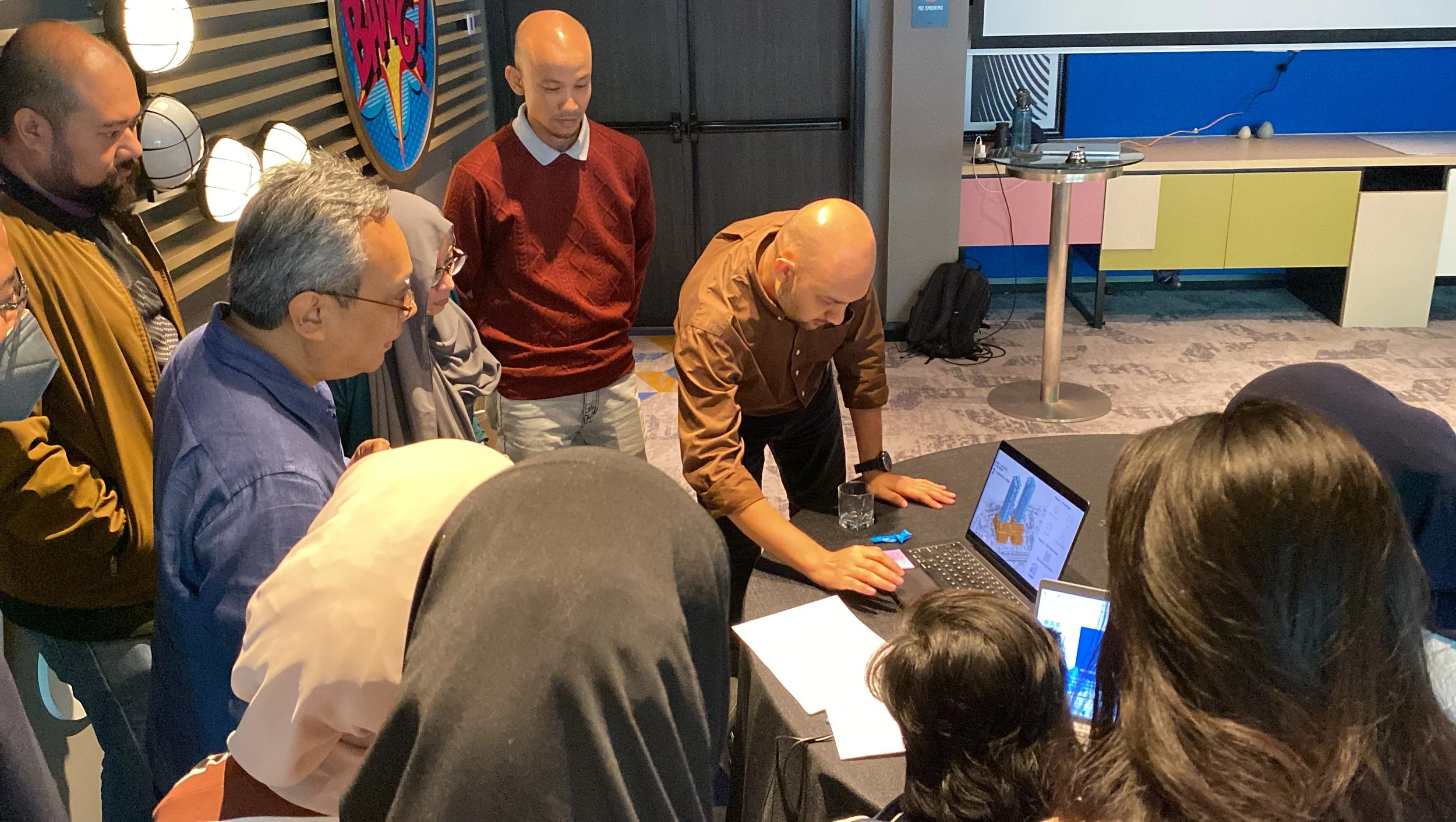 A group of diverse people gathered around a table, observing a man demonstrating something on a laptop in a vibrant, modern room with colorful wall decor and bright lighting.