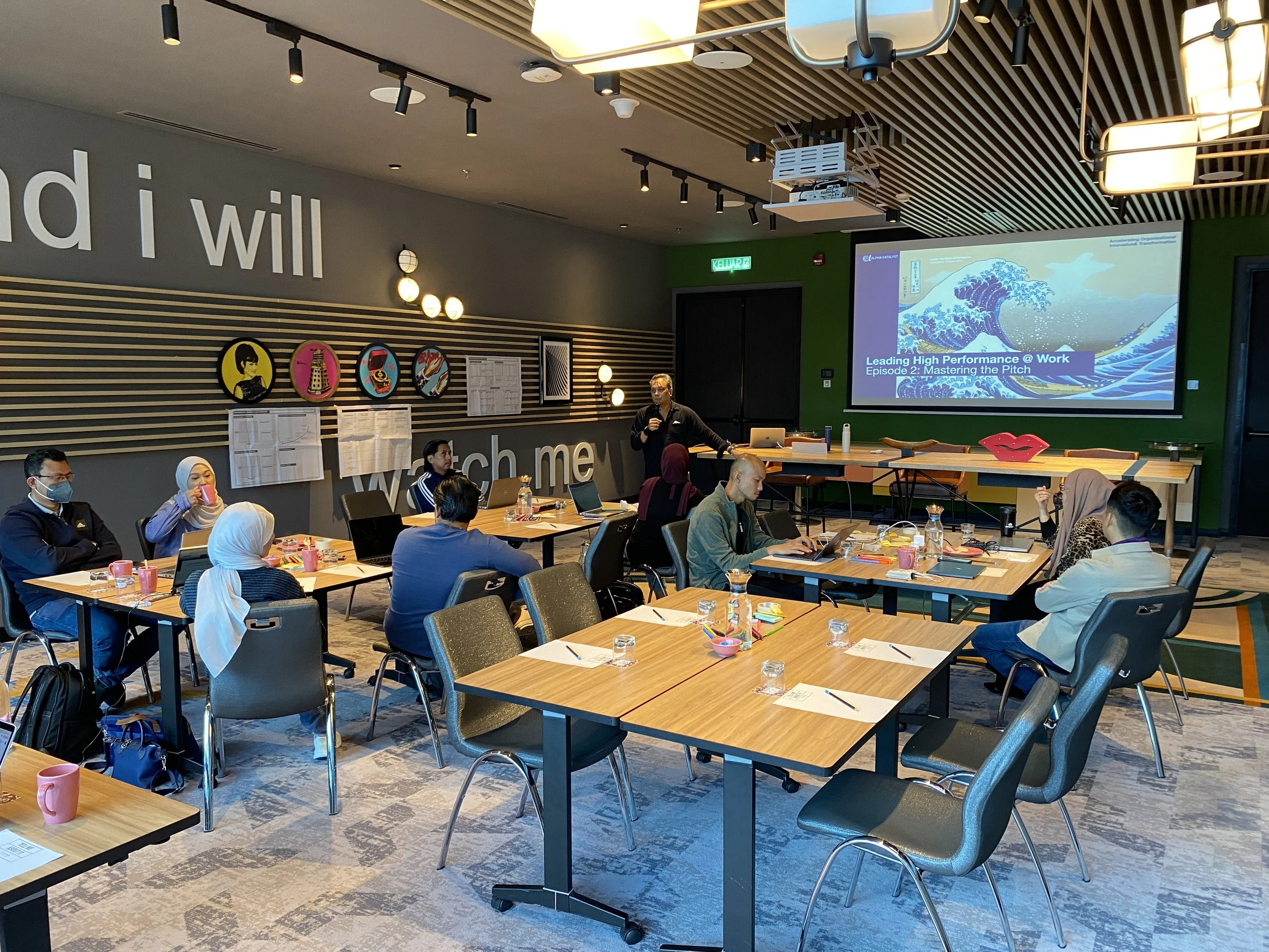 A conference room with people attending a presentation. There is a man with a microphone at the front, a large screen displaying a slide titled 'Leading High Performance @ Work,' and various artworks on the wall.