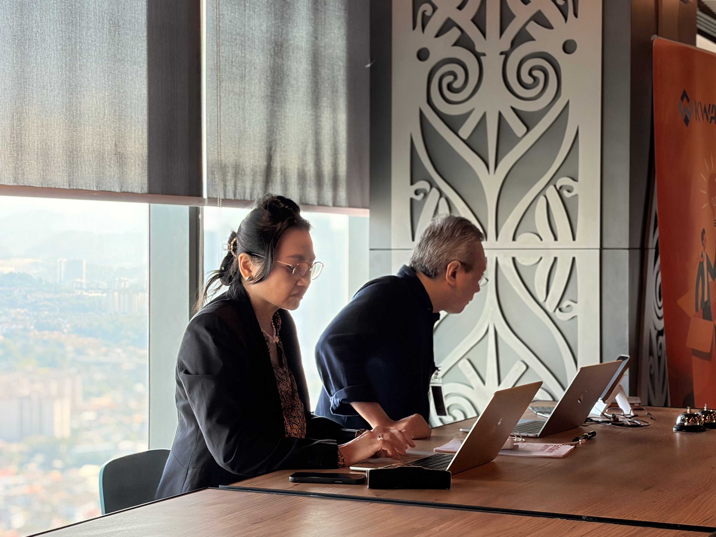 Three individuals sitting at a conference table, working on laptops, in a modern office with large windows and decorative wall panels.