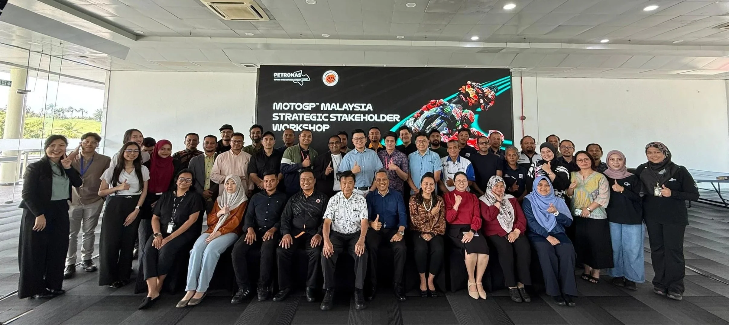 Group of people posing in front of a large screen at the MOTOGP Malaysia Strategic Stakeholder Workshop, with some individuals giving thumbs-up and smiling.