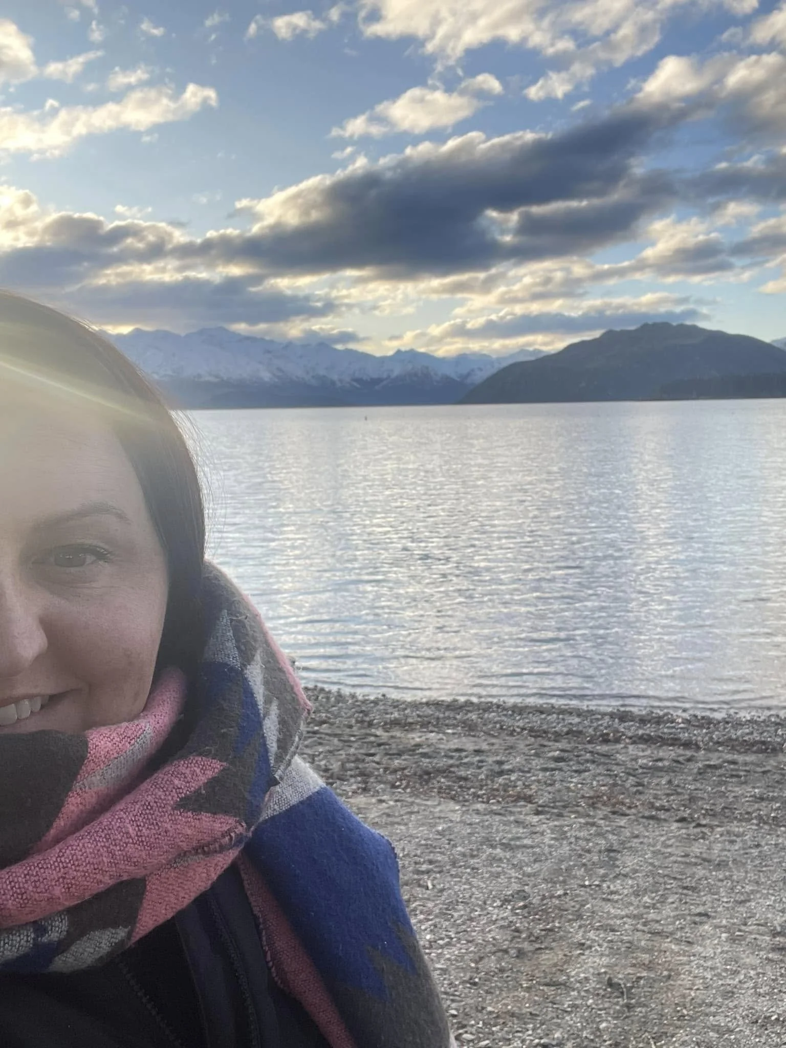 Partial view of a person's face and shoulder in the foreground, wearing outdoor clothing, with a body of water, mountains, and a cloudy sky in the background.