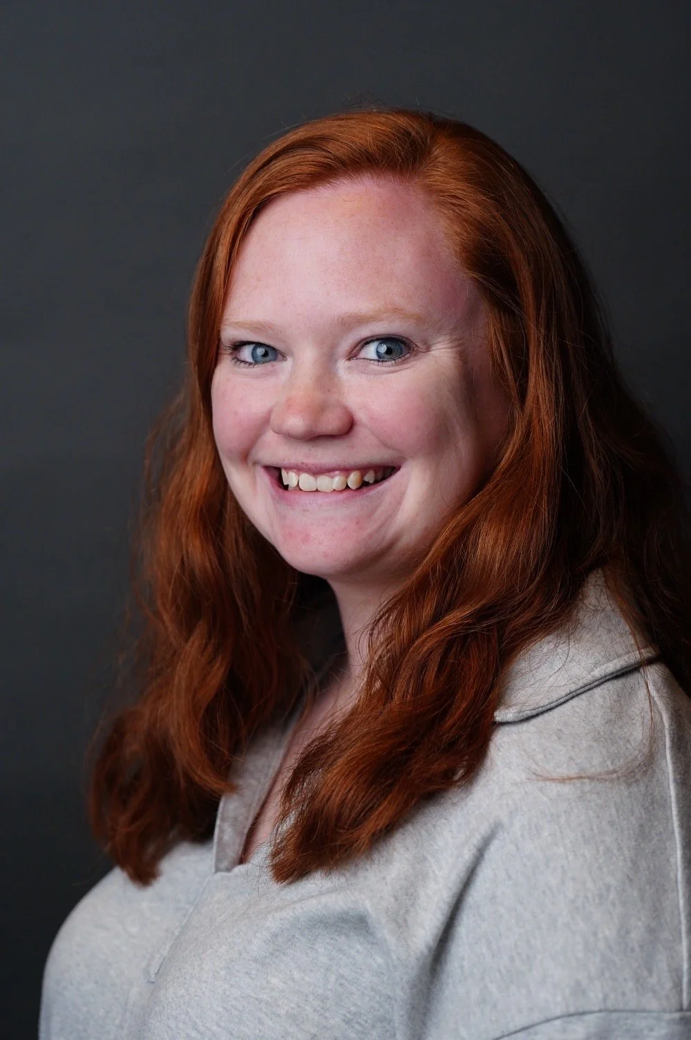 Portrait of a smiling woman with long red hair, blue eyes, and a light-colored top, against a dark background.