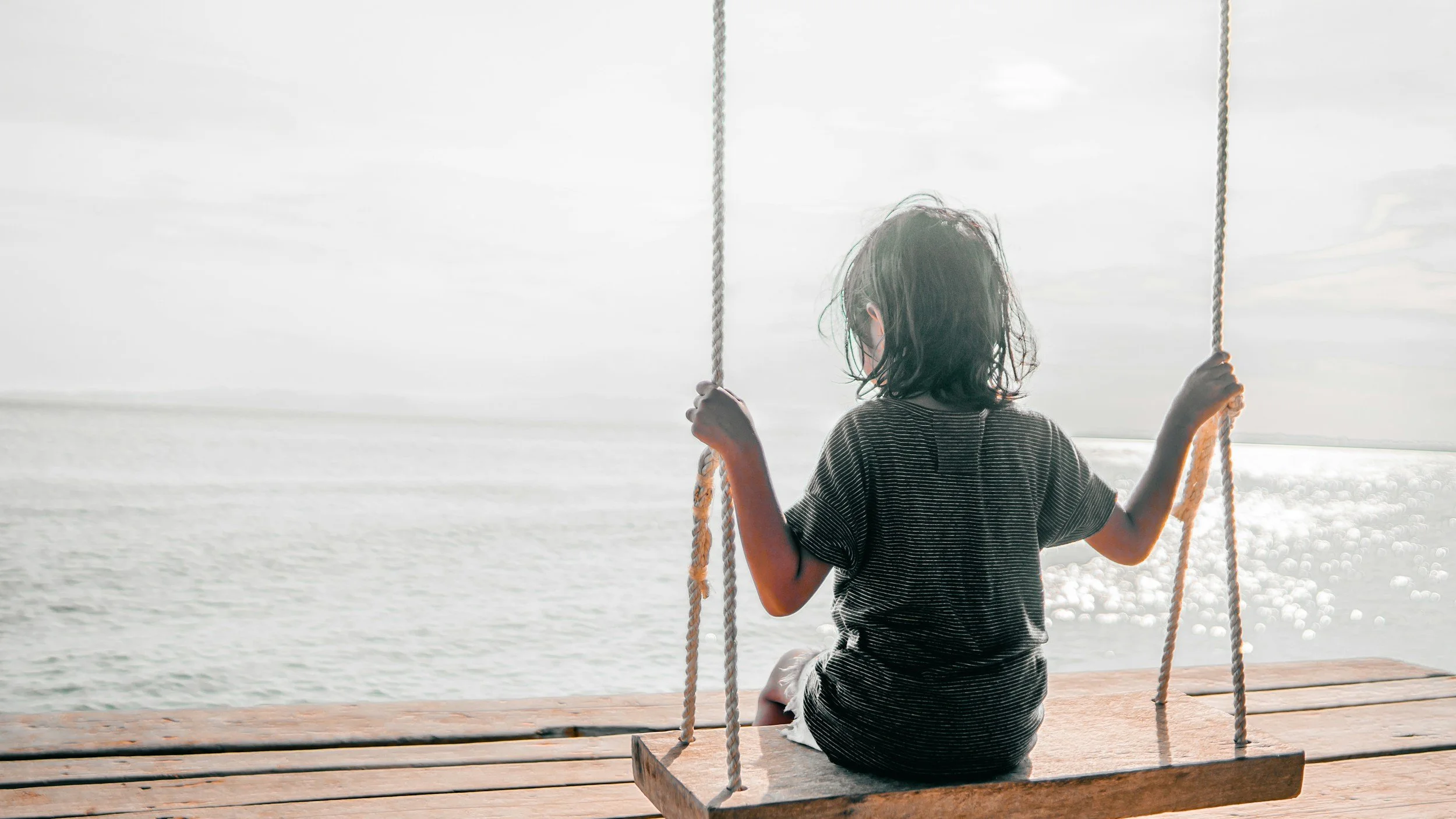 Child sitting on a wooden swing at the beach, facing the water, with overcast sky.