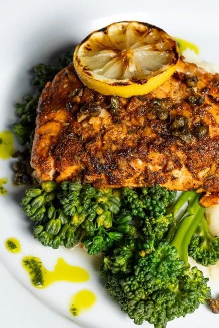 Cooked fish fillet topped with grilled lemon slice, served with broccoli florets and drizzled with yellow sauce on a white plate.