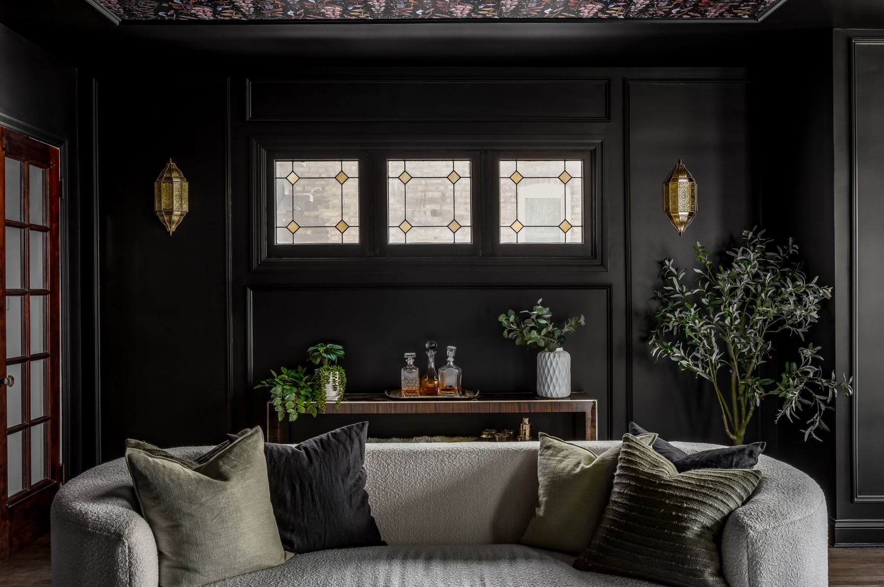 A cozy living room corner with black walls, a beige curved sofa with green and black pillows, a wooden sideboard with glass decanters, a tall leafy plant, and three small windows with decorative glass panes.