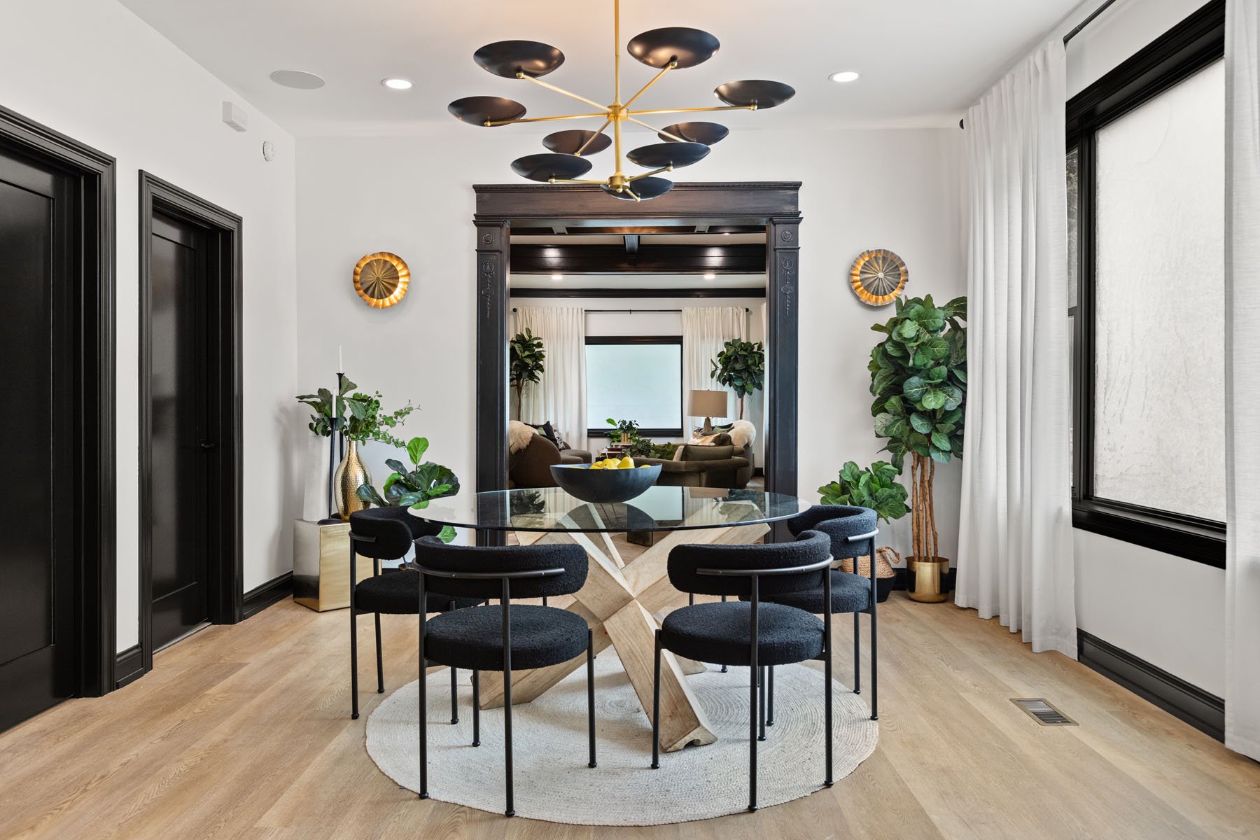 Dining room with modern black chairs around a glass table on a round rug, decorated with green plants, gold accents, black doors, and large windows with white curtains, leading to a living room with sofas and a large window.