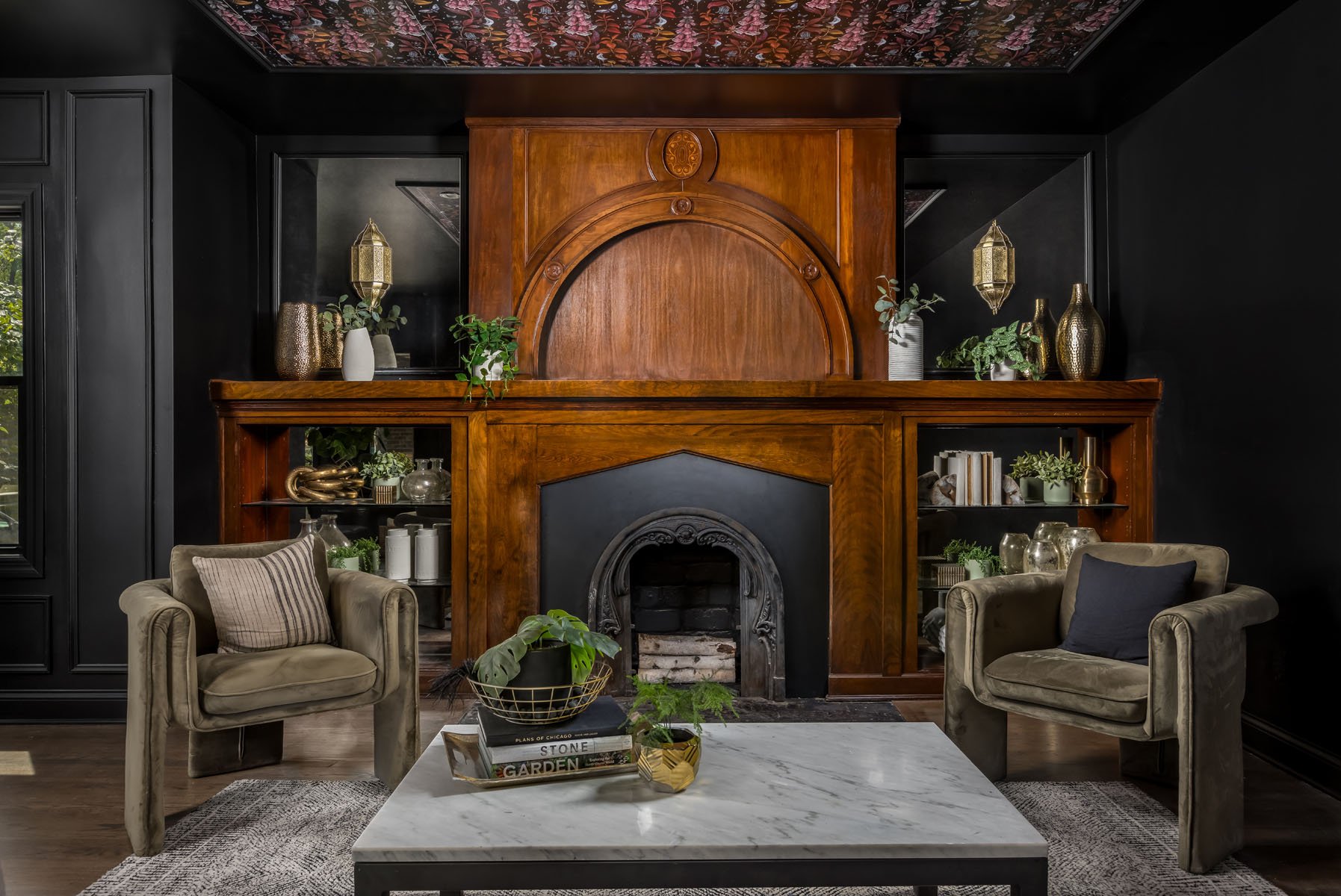 Living room with a black and wood fireplace surrounded by black walls, decorated with green potted plants and gold vases. Two beige armchairs with pillows, a coffee table with plants and books, and a patterned ceiling.