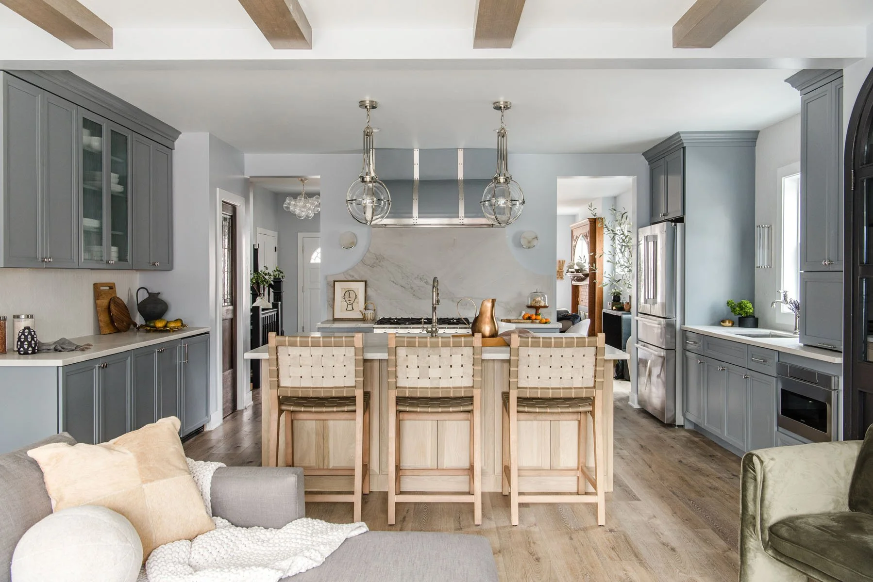 A modern kitchen with gray cabinets, a white marble backsplash, and a central island with four wooden barstools. The ceiling features exposed wooden beams, and there are pendant lights hanging above the island. The room has hardwood floors, some deco