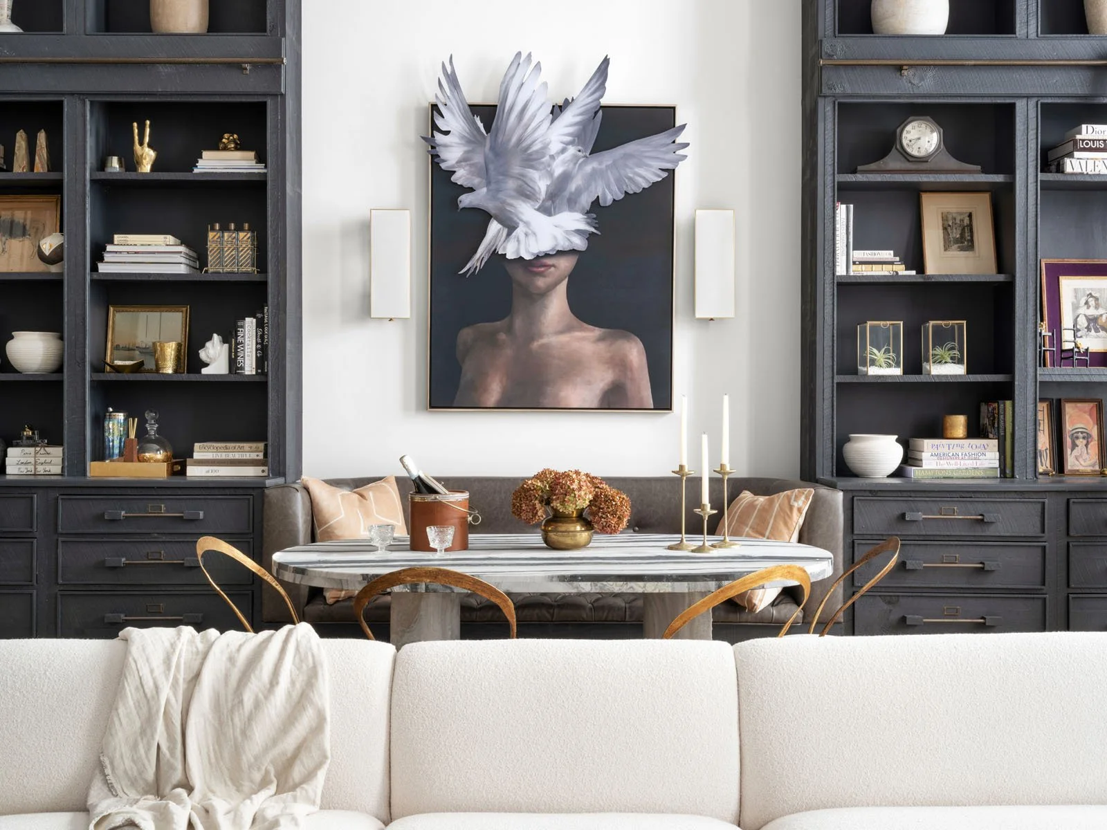 A living room with dark built-in bookshelves filled with decor, books, and framed photos. A large painting of a woman with a dove over her eyes is centered above a leather banquette, behind a dining table. On the table are candles, flowers, and glass