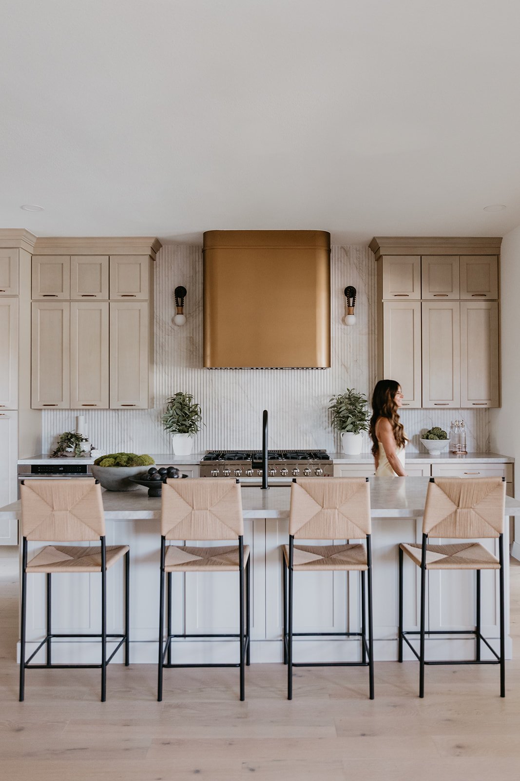 Modern kitchen with beige cabinetry, a large white island with four bar stools, a gold range hood, and a woman with long brown hair standing at the counter.