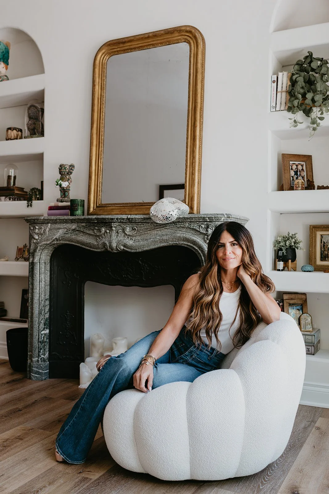 A woman with long wavy hair sitting on a white, textured armchair in a living room with a marble fireplace, a gold-framed mirror, and decorative shelves with books and plants.