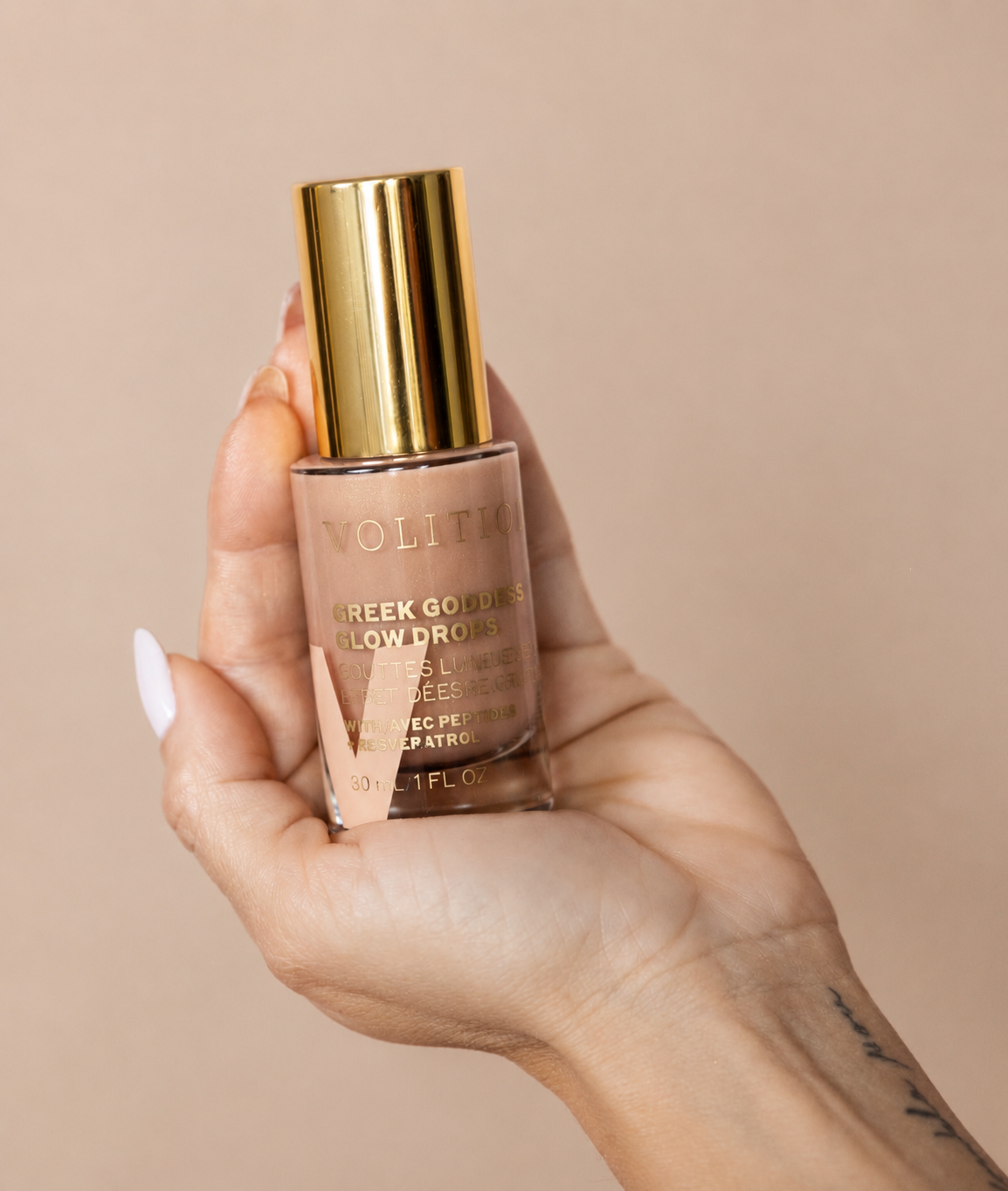 A hand holding a bottle of Volition Greek Goddess Glow Drops, a skincare product with a gold cap and a light pink liquid inside.