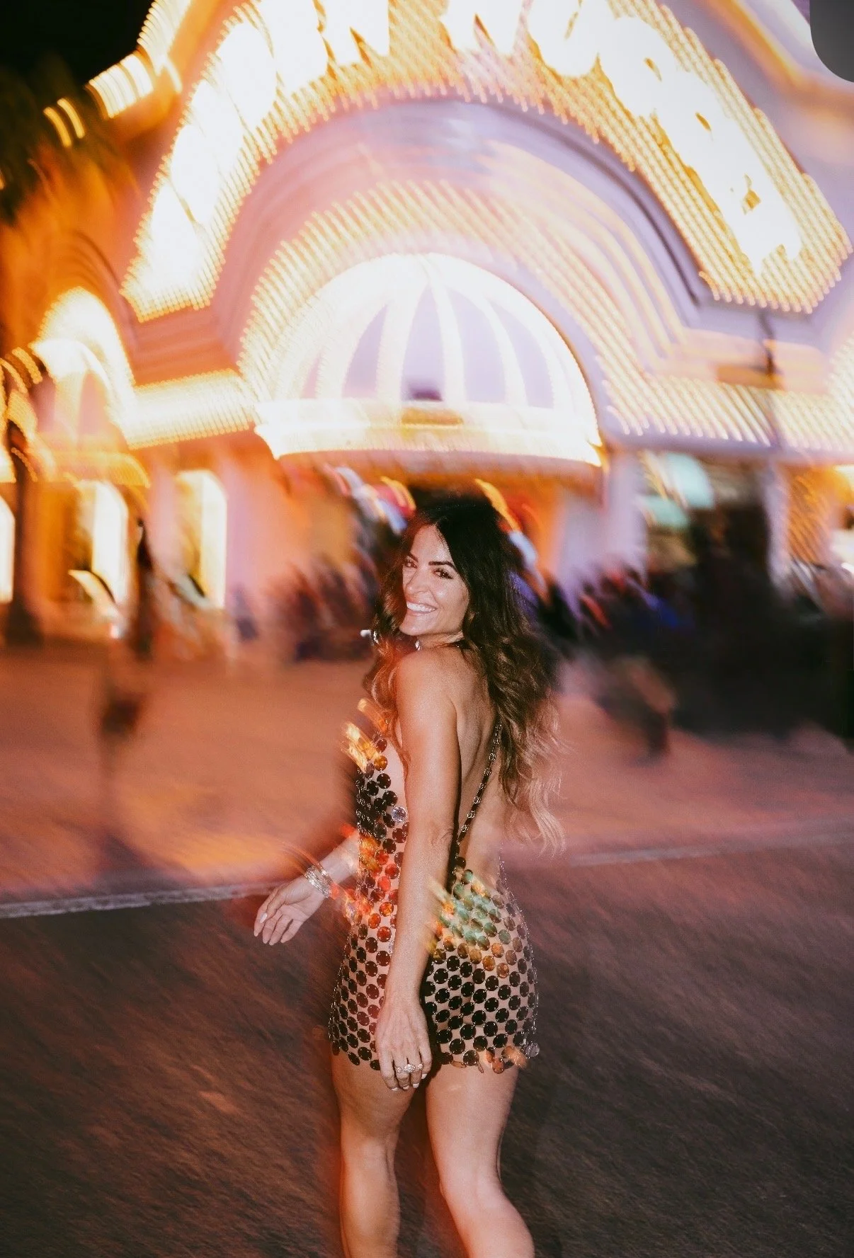 A woman smiling and walking in front of a brightly lit carnival or fair at night, with colorful lights and blurred figures in the background.