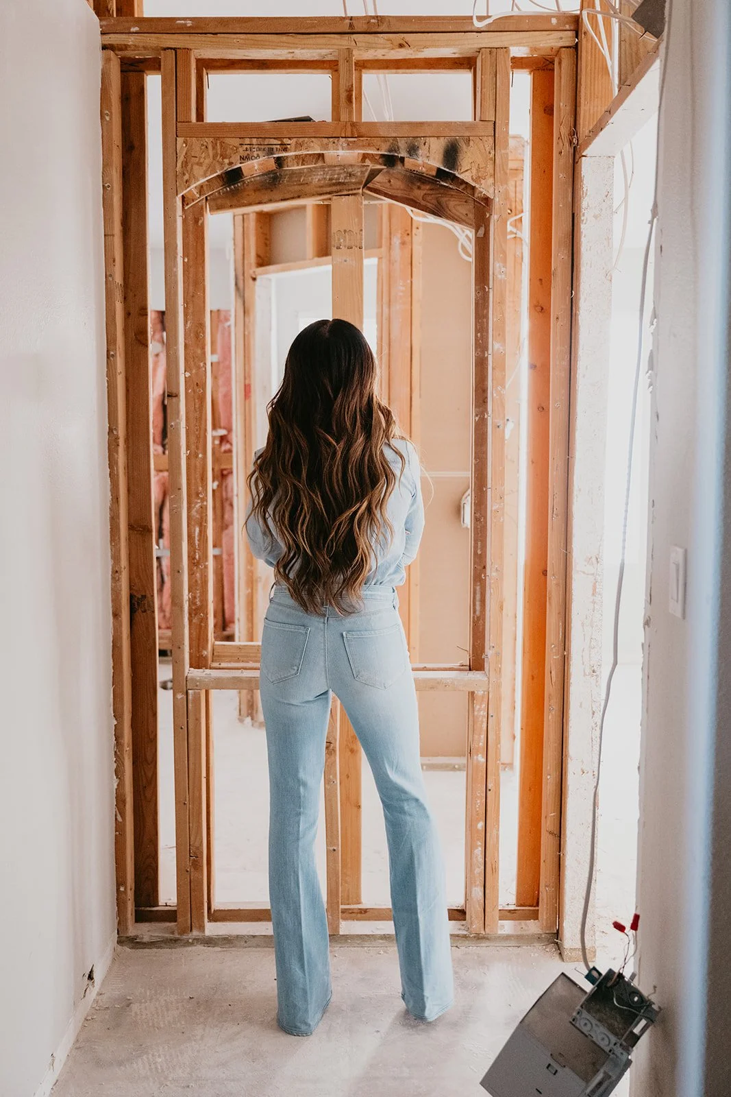 A woman with long wavy hair wearing light blue jeans and a denim jacket standing in front of a partially built wooden wall inside a house under renovation.