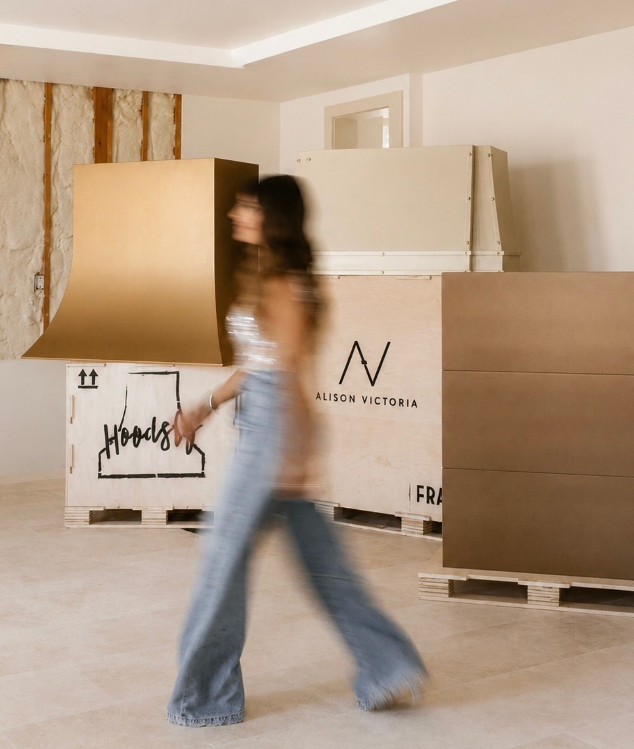 A woman in a sleeveless top and jeans walking past wooden crates in a showroom or gallery space. The crates have branding for 'Alison Victoria' and 'Hood's' with a floorplan diagram.