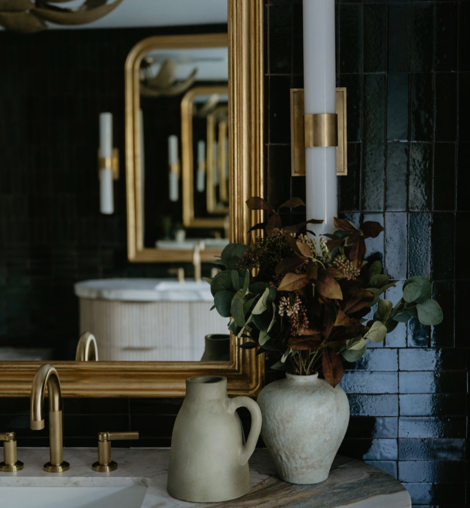 A bathroom with black tiled walls, a large mirror with a gold frame, a white vessel sink, a gold faucet, and two decorative vases with flowers and greenery on a marble countertop.