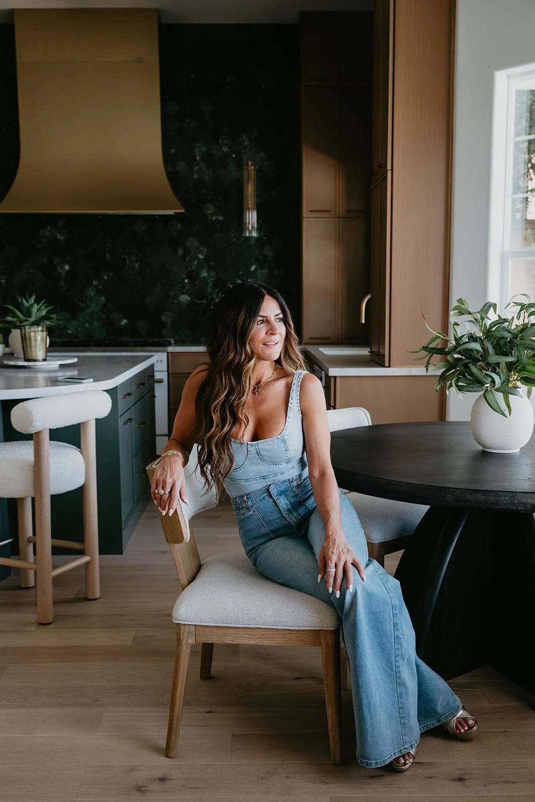 Woman sitting at a round table in a modern kitchen with wooden cabinetry, a black marble backsplash, and a large window. She has long wavy hair and is wearing a blue denim top and jeans.
