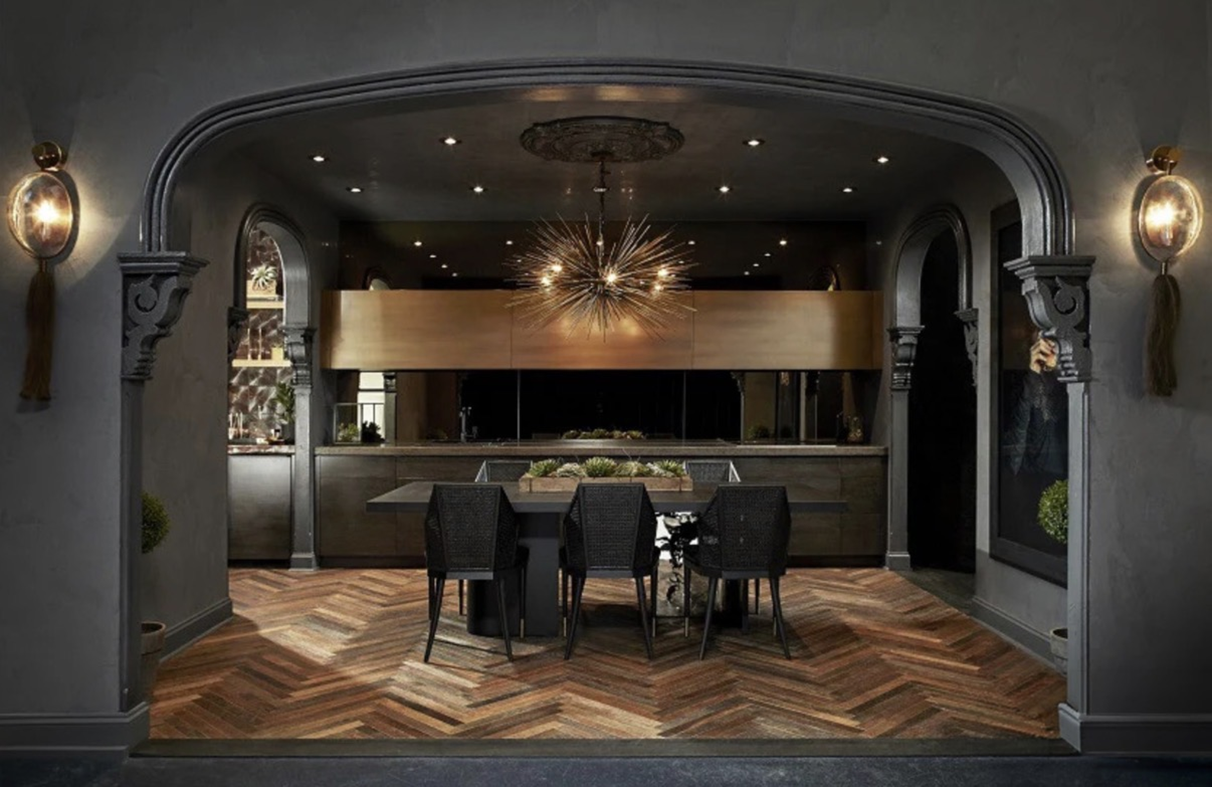 An elegant dining space with a dark gray wall, ornate wall sconces, wooden archways, and a dark wooden dining table with black chairs. A modern chandelier hangs above the table, and the wood flooring has a herringbone pattern.