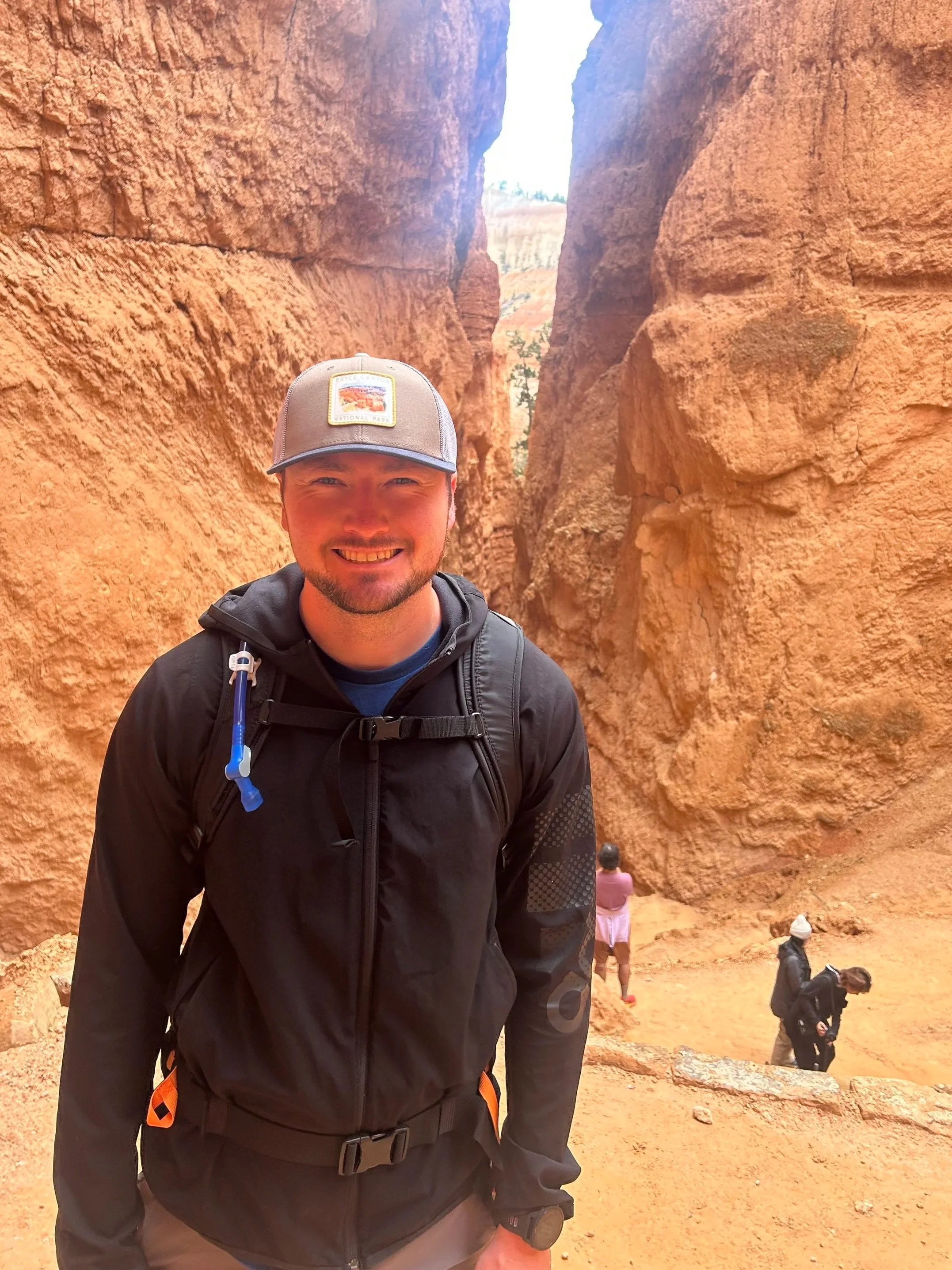 Man smiling in front of red rock canyon walls with several other people in the background.