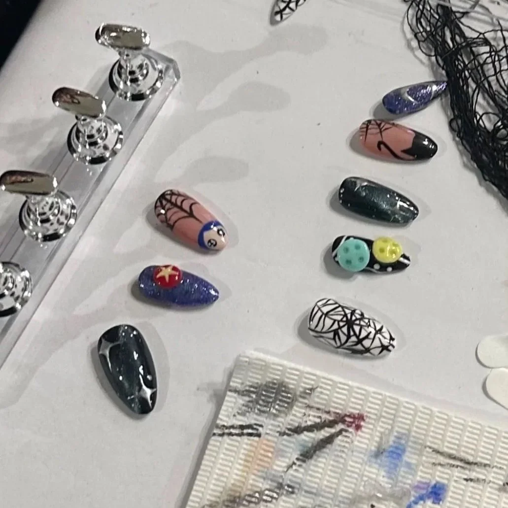 Collection of decorated nail art tips arranged on a white surface, with some featuring abstract designs, spider web patterns, and colorful accents, alongside metal objects and a mesh fabric.