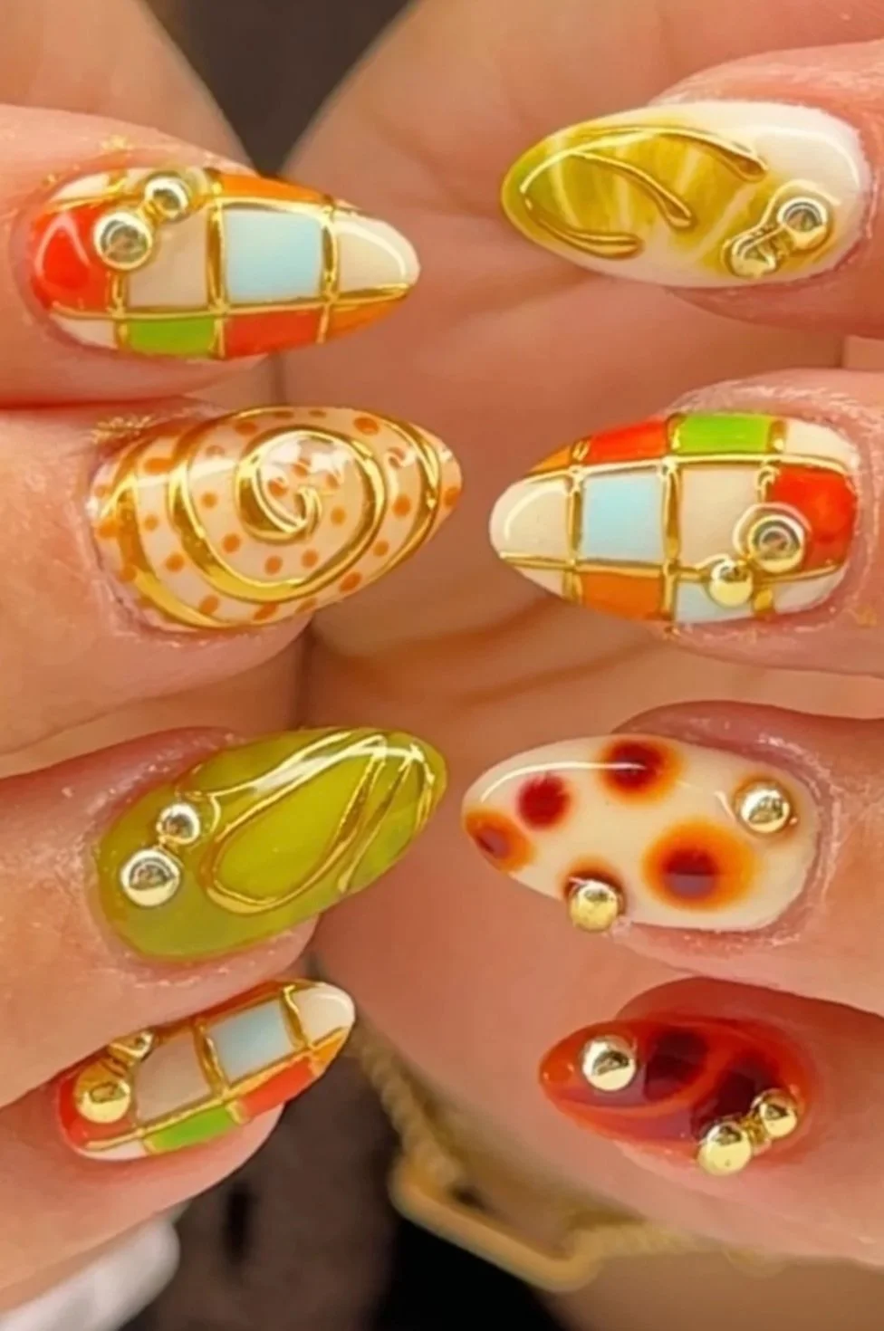 Close-up of a person's fingernails with colorful nail art featuring various patterns, colors, and gold accents.