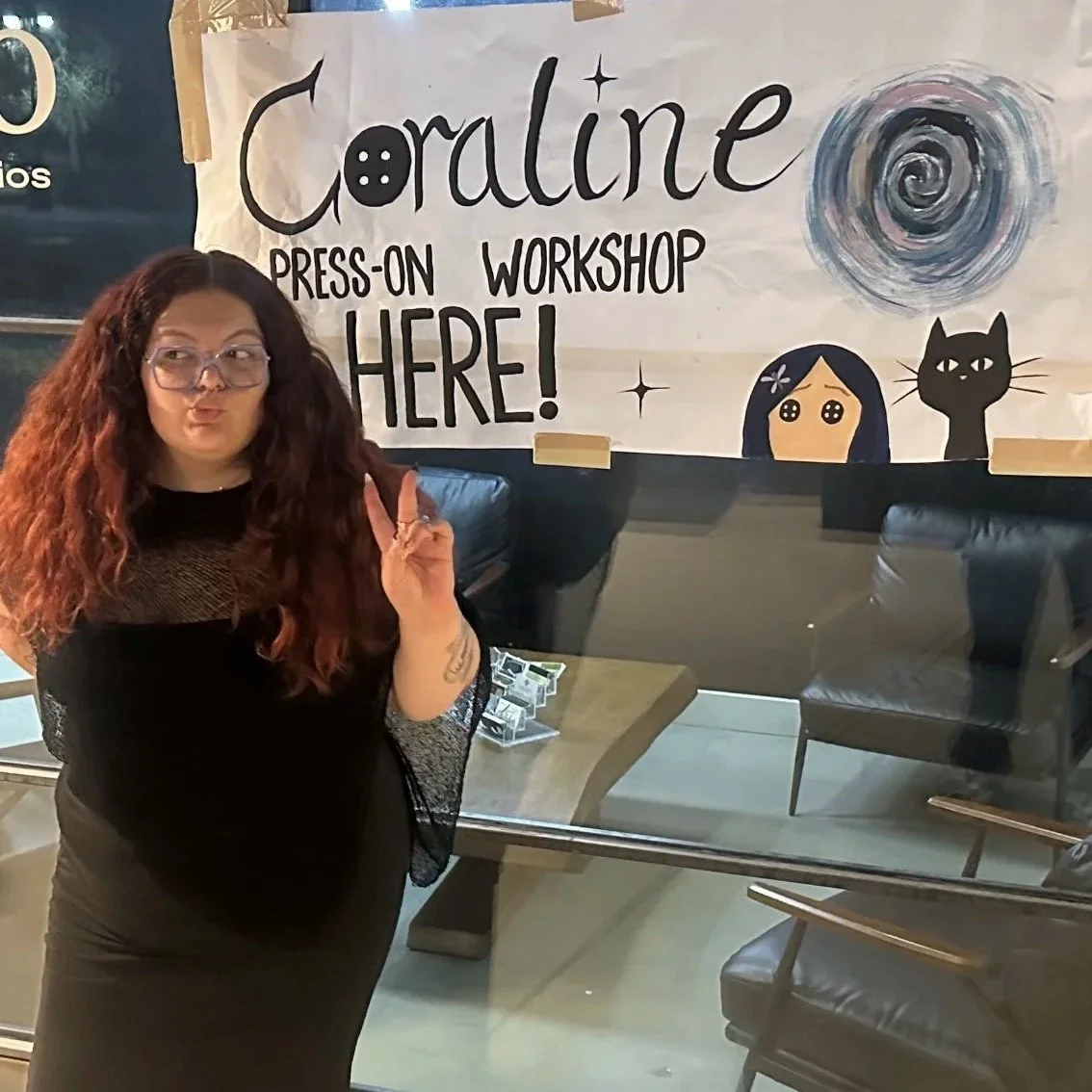 A woman with long, curly red hair and glasses making a peace sign in front of a hand-drawn sign that reads 'Coraline Press-On Workshop Here!' with art of Coraline and a black cat.