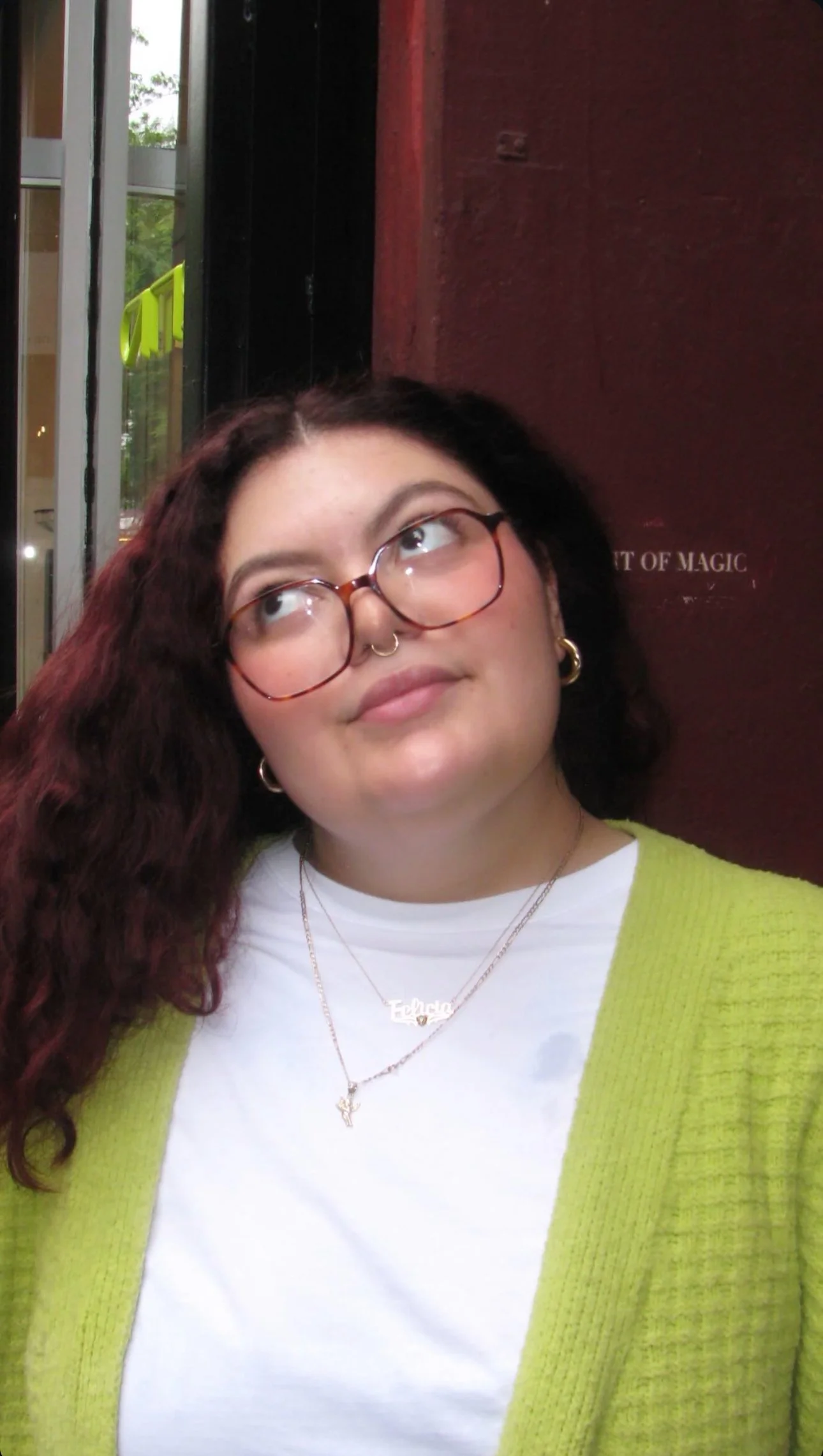 A young woman with curly dark hair, wearing glasses, a white shirt, and a lime green sweater. She has layered necklaces and small hoop earrings. She is looking up with a thoughtful expression, standing indoors near a window and a dark red wall.