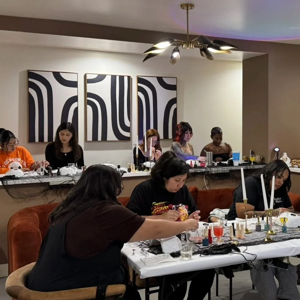 Group of women gathered around tables working on artistic projects, with various art supplies, candles, and drinks in a well-lit room with modern black and white wall art.