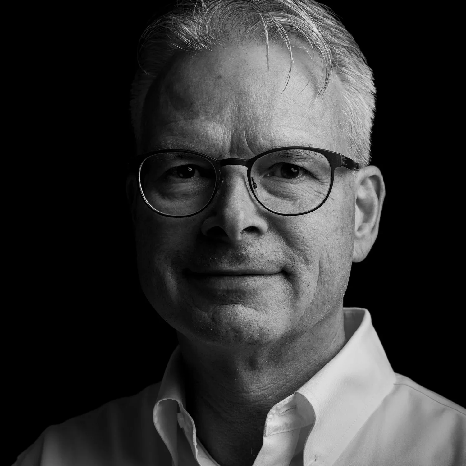 Schwarz-weiß Portrait eines älteren Mannes mit kurzen, grauen Haaren, Brille und weißem Hemd, vor schwarzem Hintergrund.