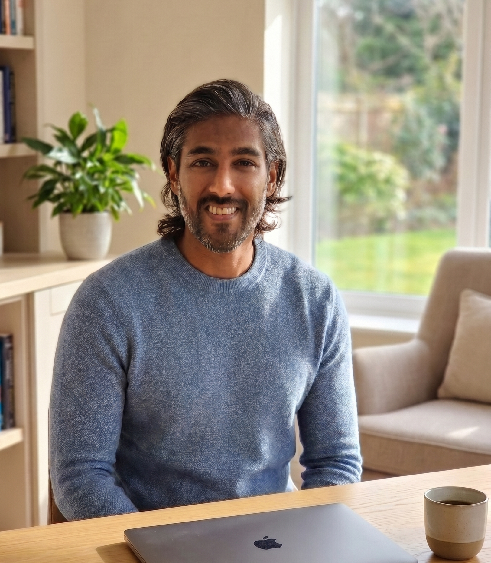 A man with dark hair and a beard, wearing a gray sweater, is sitting at a wooden table in a room with a large window showing greenery outside. There is a laptop and a cup of coffee on the table, and a potted plant and bookshelf are visible in the background.