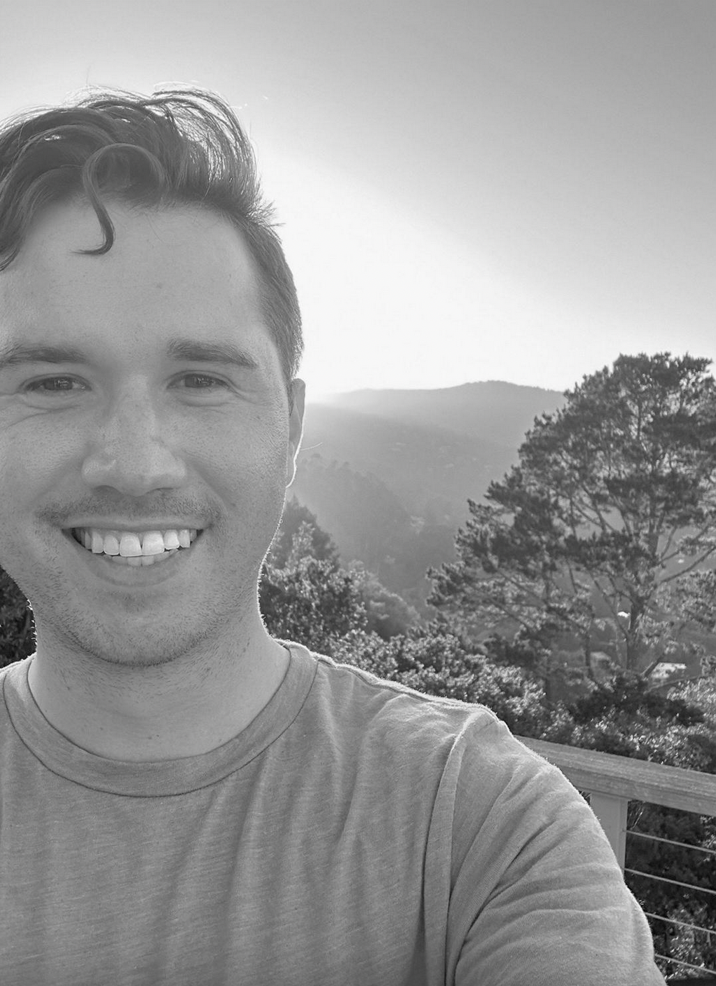 A young man smiling for a selfie outdoors, with trees and mountains in the background, black and white photo.