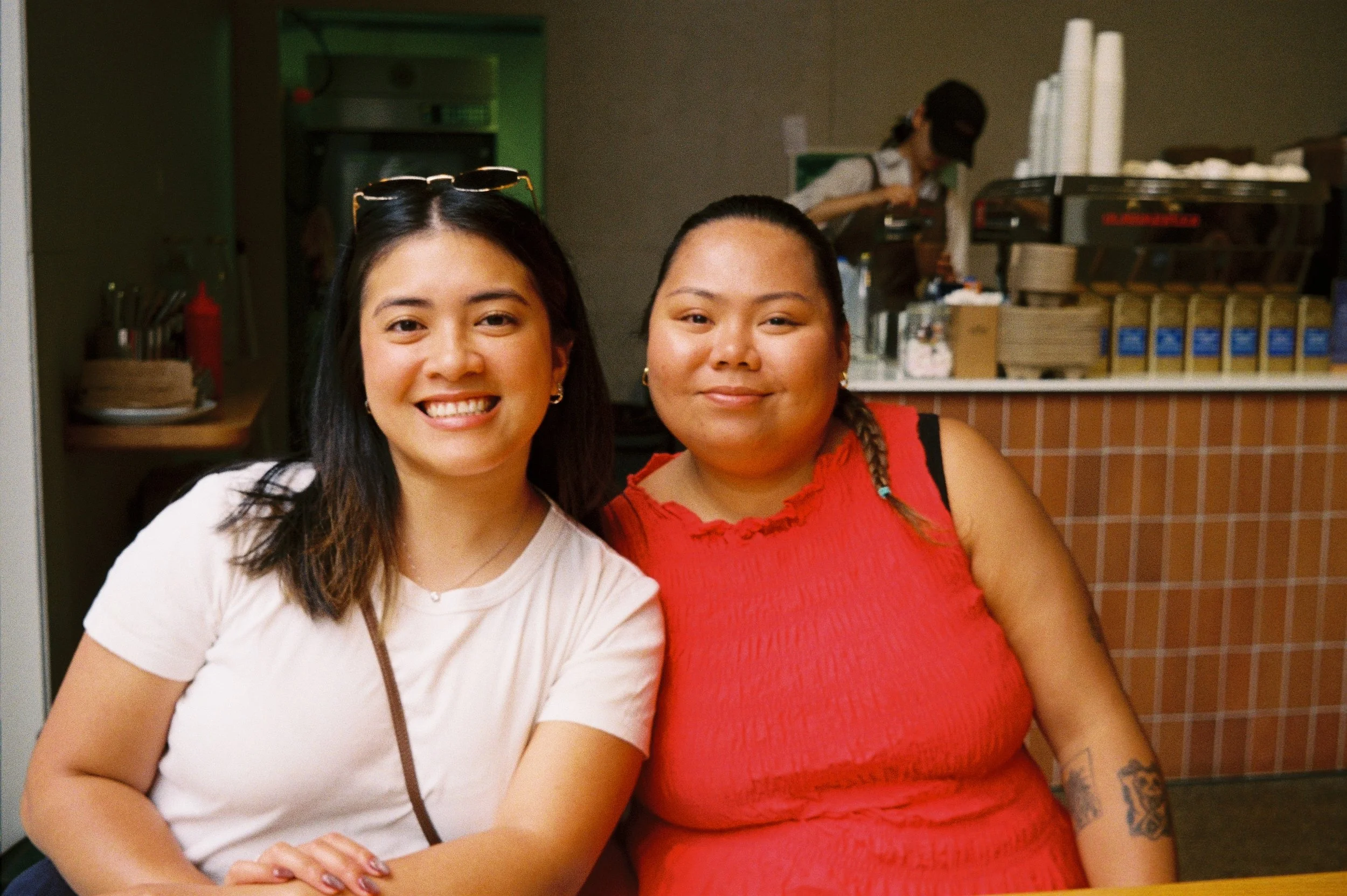 Two smiling women sitting close together at a counter in a restaurant, with a food preparation area and a woman working in the background.