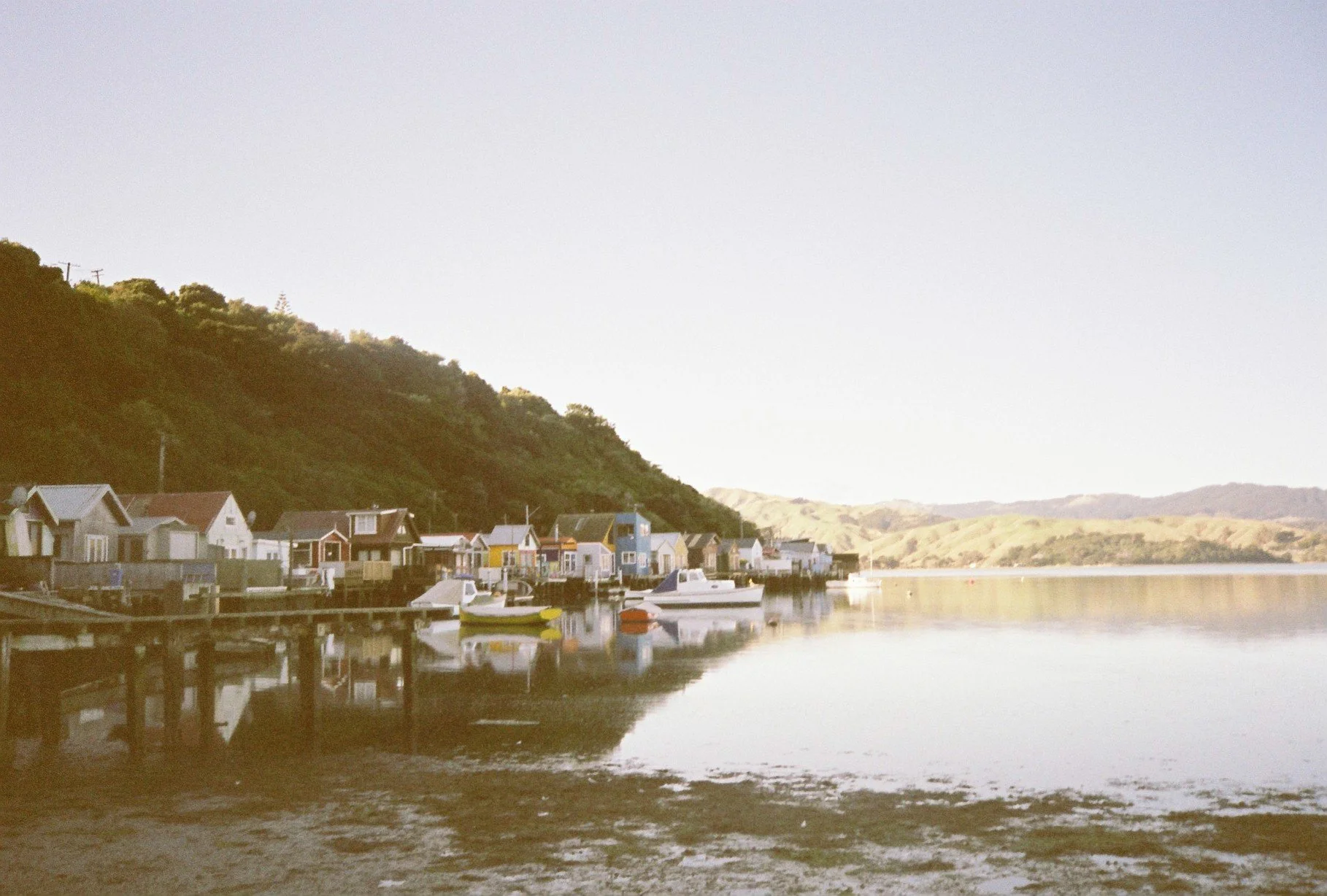 A small harbor with colorful houses on stilts along the waterfront, boats floating on calm water, and a green hillside in the background under a light sky.