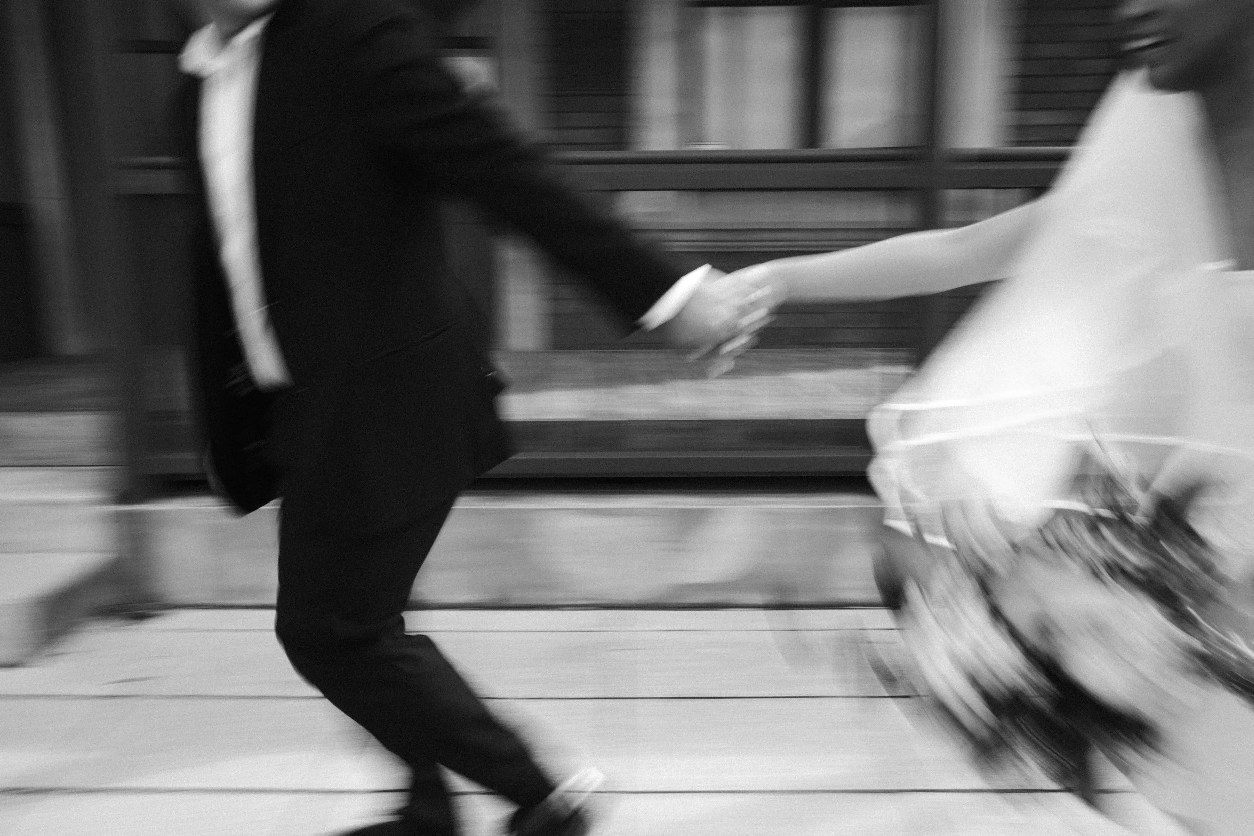 Blurred black and white photo of two people holding hands while walking, one wearing a dark jacket and the other a light dress.