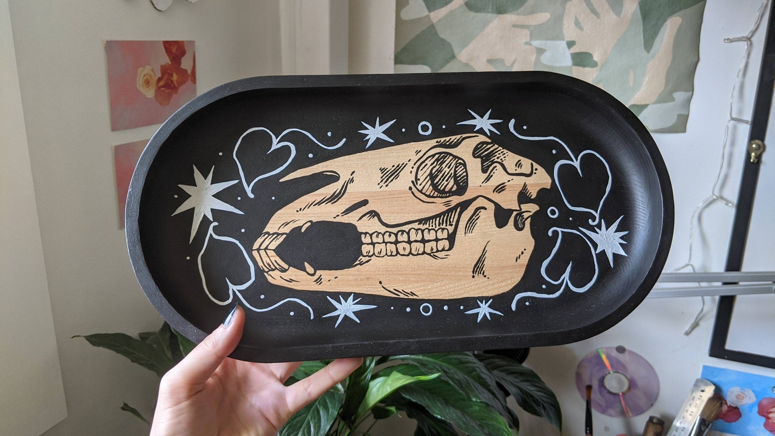 A black oval tray featuring a design of a detailed human skull with an open mouth, surrounded by white stars, heart shapes, and swirling lines, held by a person's hand. In the background, there are pink and red floral wall art and various objects on 