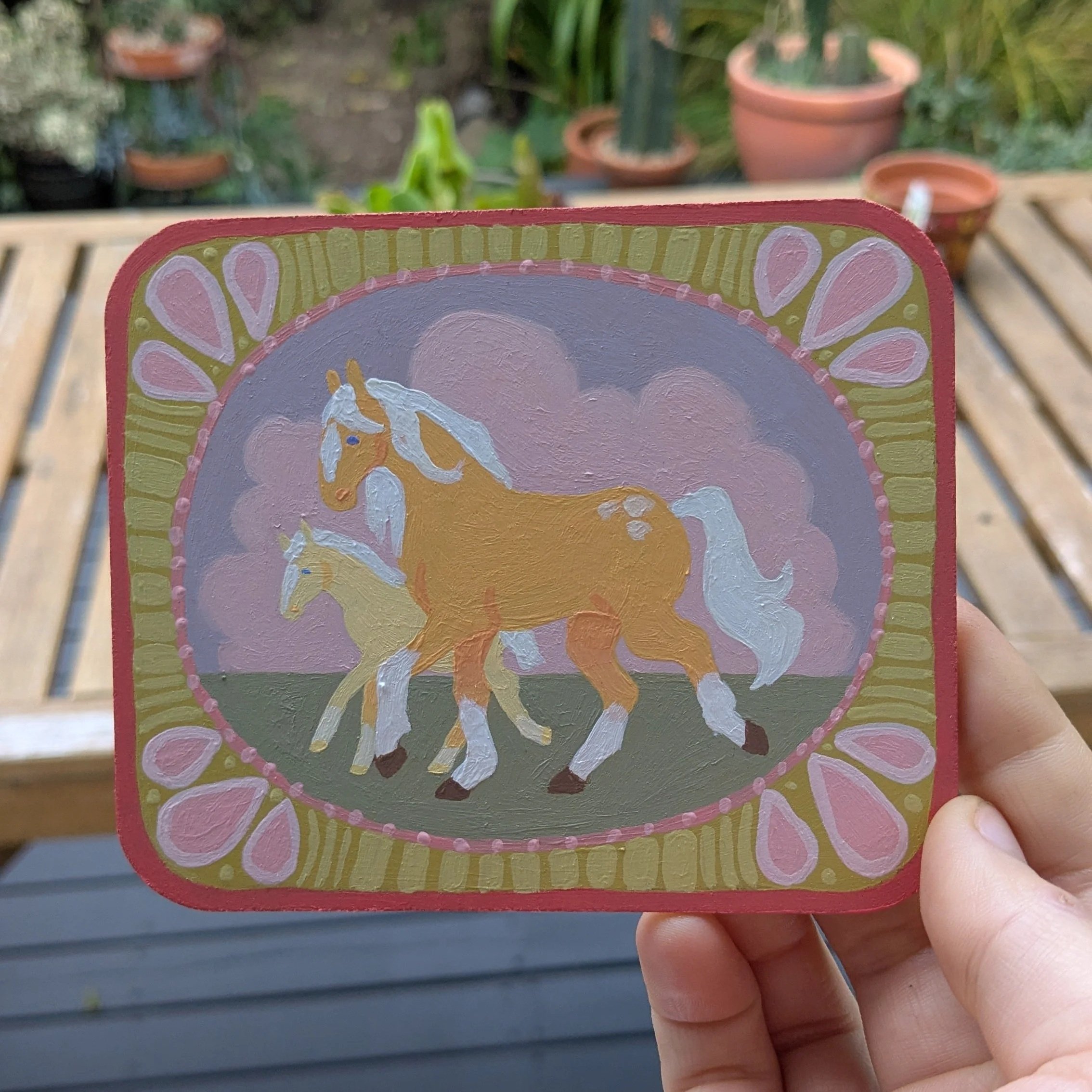 A person holding a small colorful wooden sign with a unicorn design, outdoor background with potted plants.