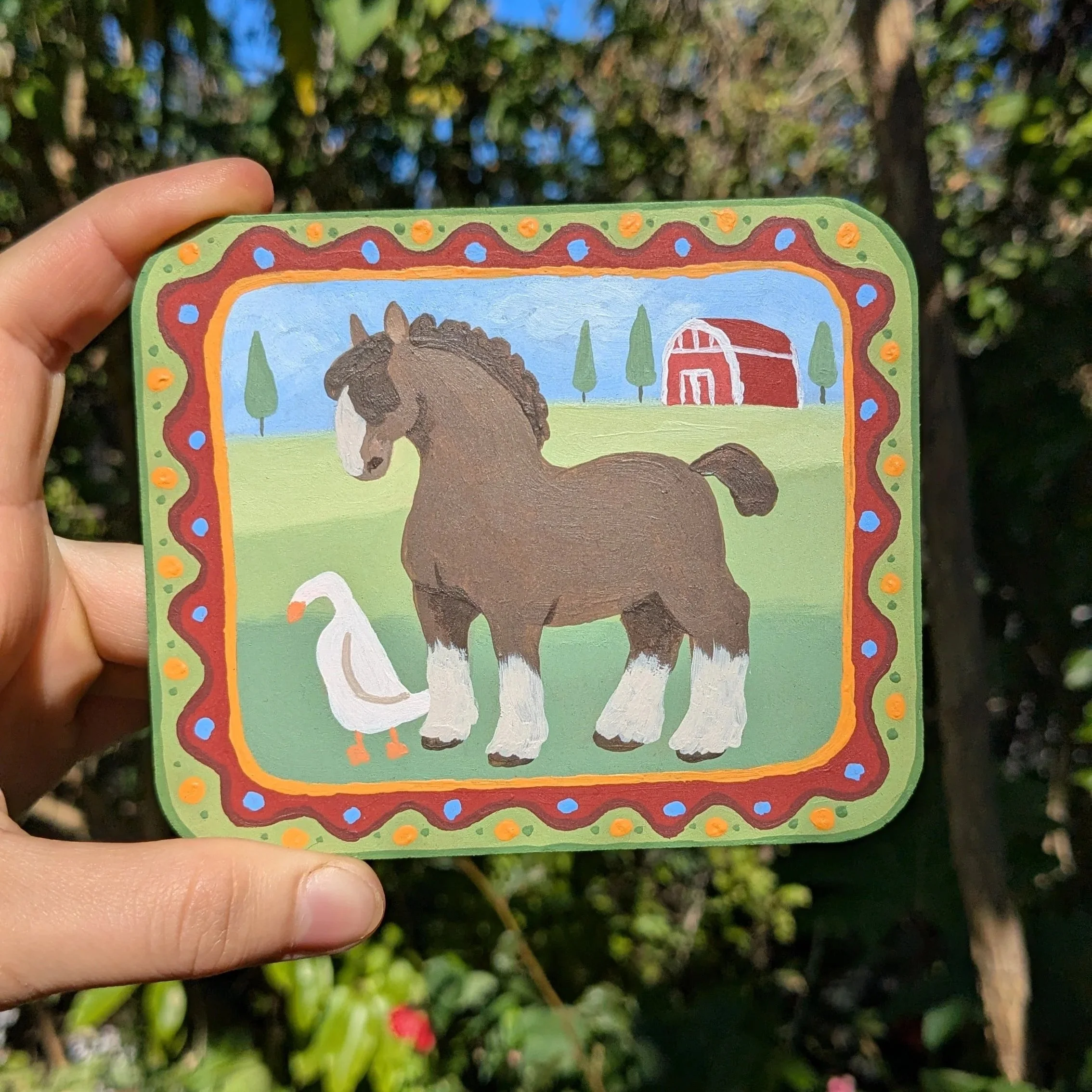 Colorful illustrated card with a horse, barn, and trees outdoors, held in a person's hand against a background of green bushes and blue sky.