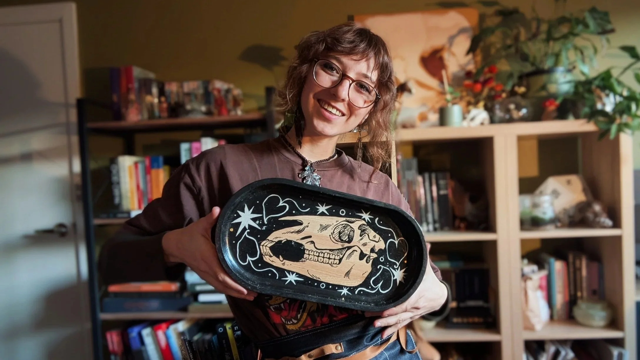 A woman with glasses and wavy hair smiling, holding a decorated skull-shaped tray in a room with a bookshelf and plants in the background.