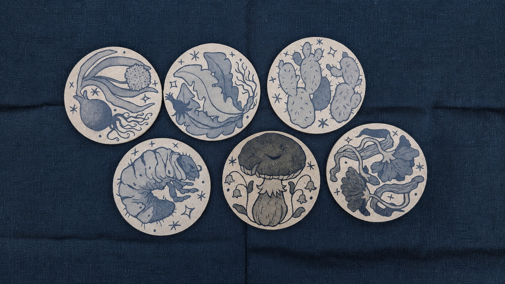 Six illustrated coasters featuring various botanical and mushroom designs arranged on a dark fabric background.