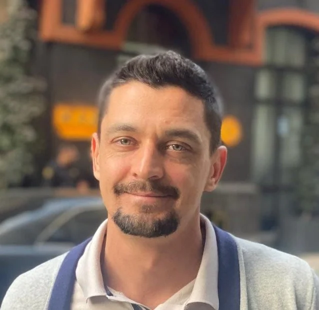 A man with short dark hair, a goatee, and a slight smile standing outdoors with a blurred urban background.