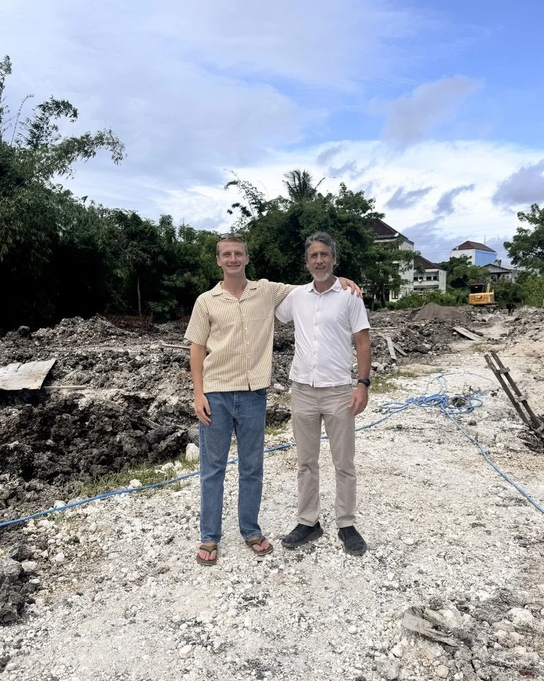 Two men standing on a construction site with scattered gravel and dirt, arm in arm, smiling, with trees and houses in the background under a partly cloudy sky.