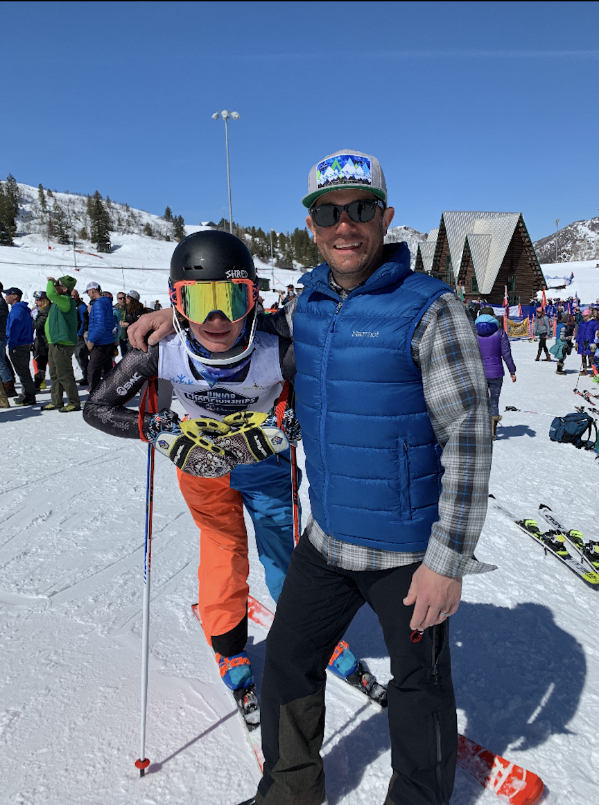 Two men in winter ski gear smiling for a photo on a snowy mountain, with skiers and ski lodges in the background.